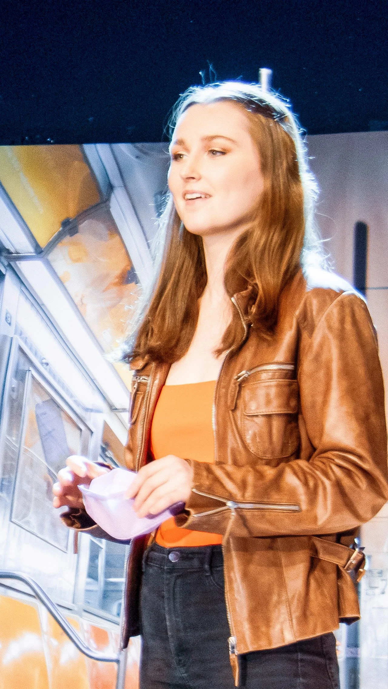 Performer in a brown leather jacket speaks onstage in Another Hundred People.