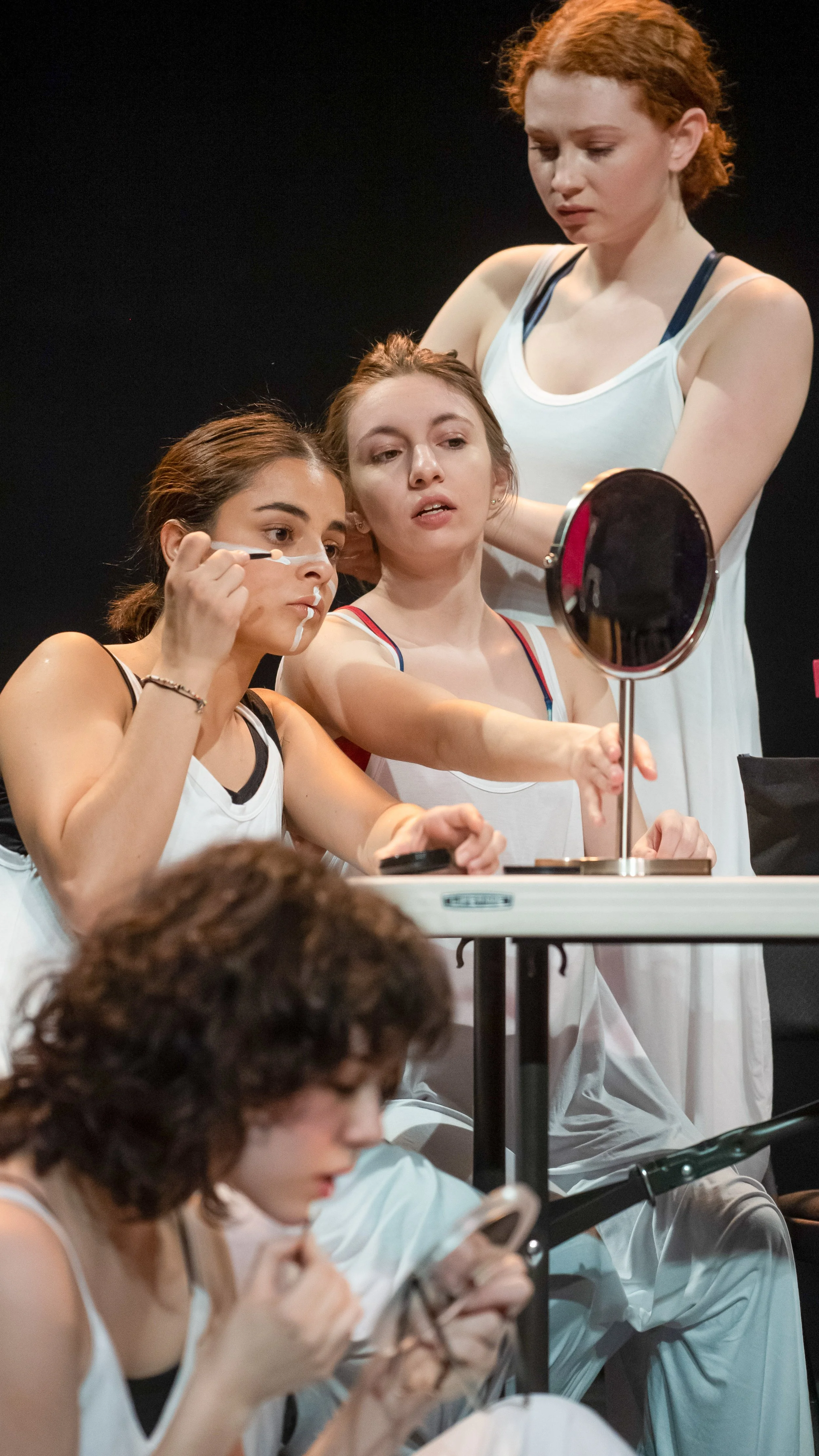 Three performers prepare at a makeup table with a handheld mirror in Dance Nation.