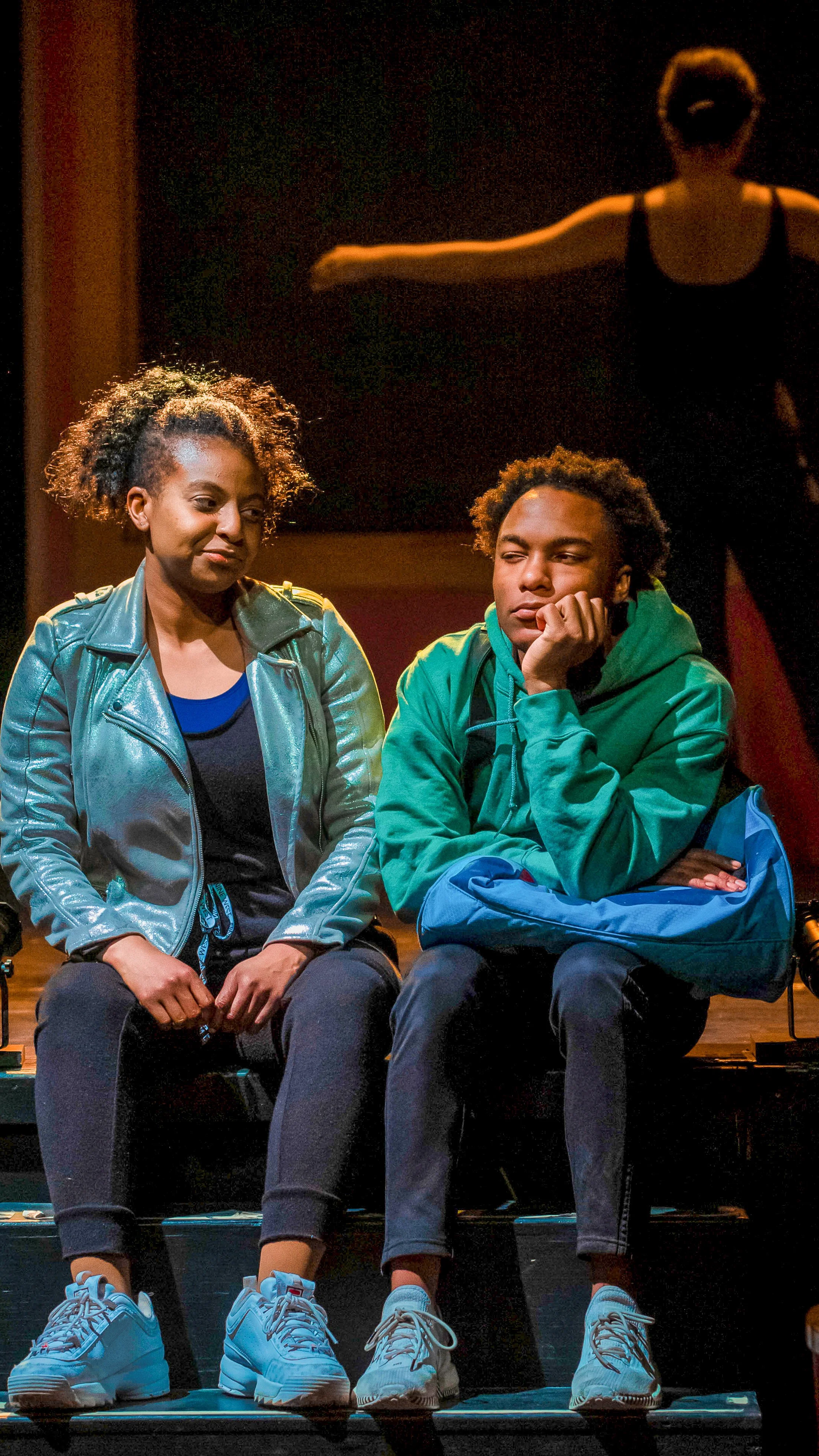 Two performers sit together on a bench, one resting her chin on her hand, in Dance Nation.