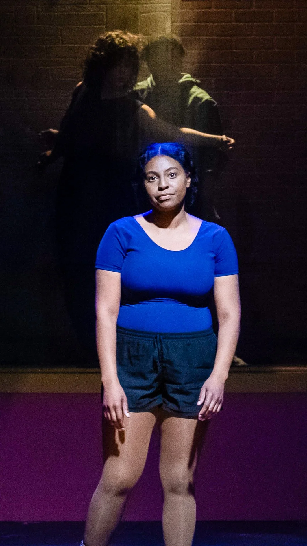 Performer stands alone center stage in a blue shirt and black shorts in Dance Nation.