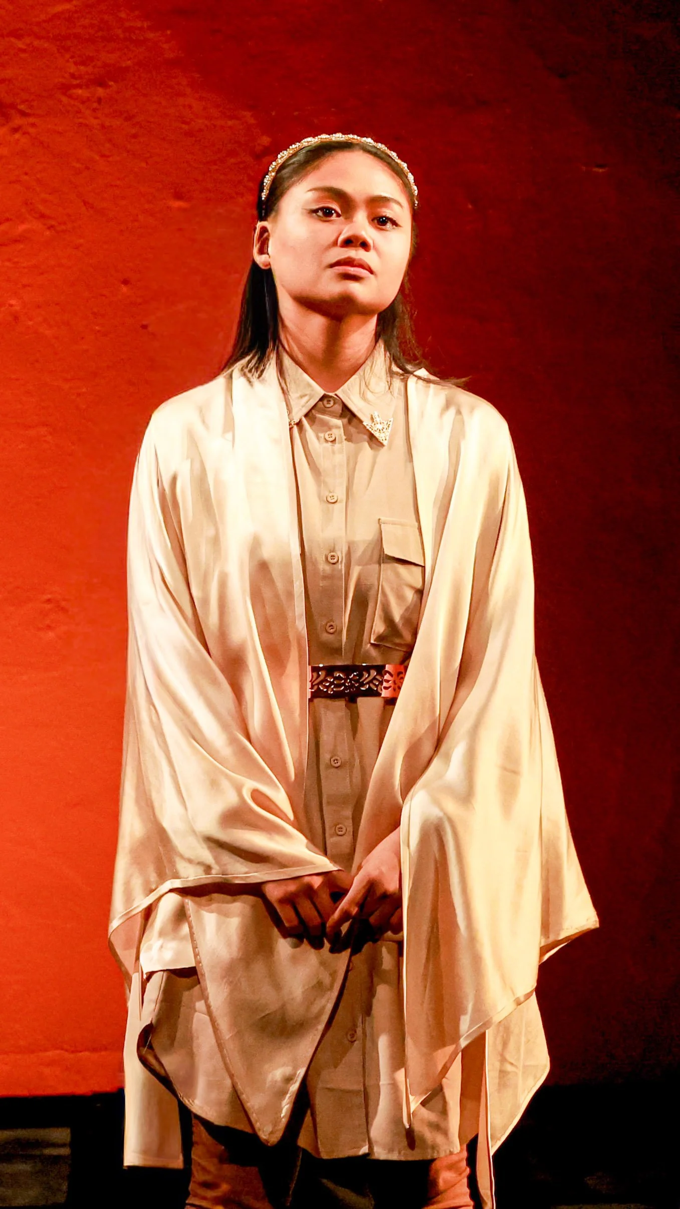 Performer in a white robe stands before a vivid red backdrop in LEAR.