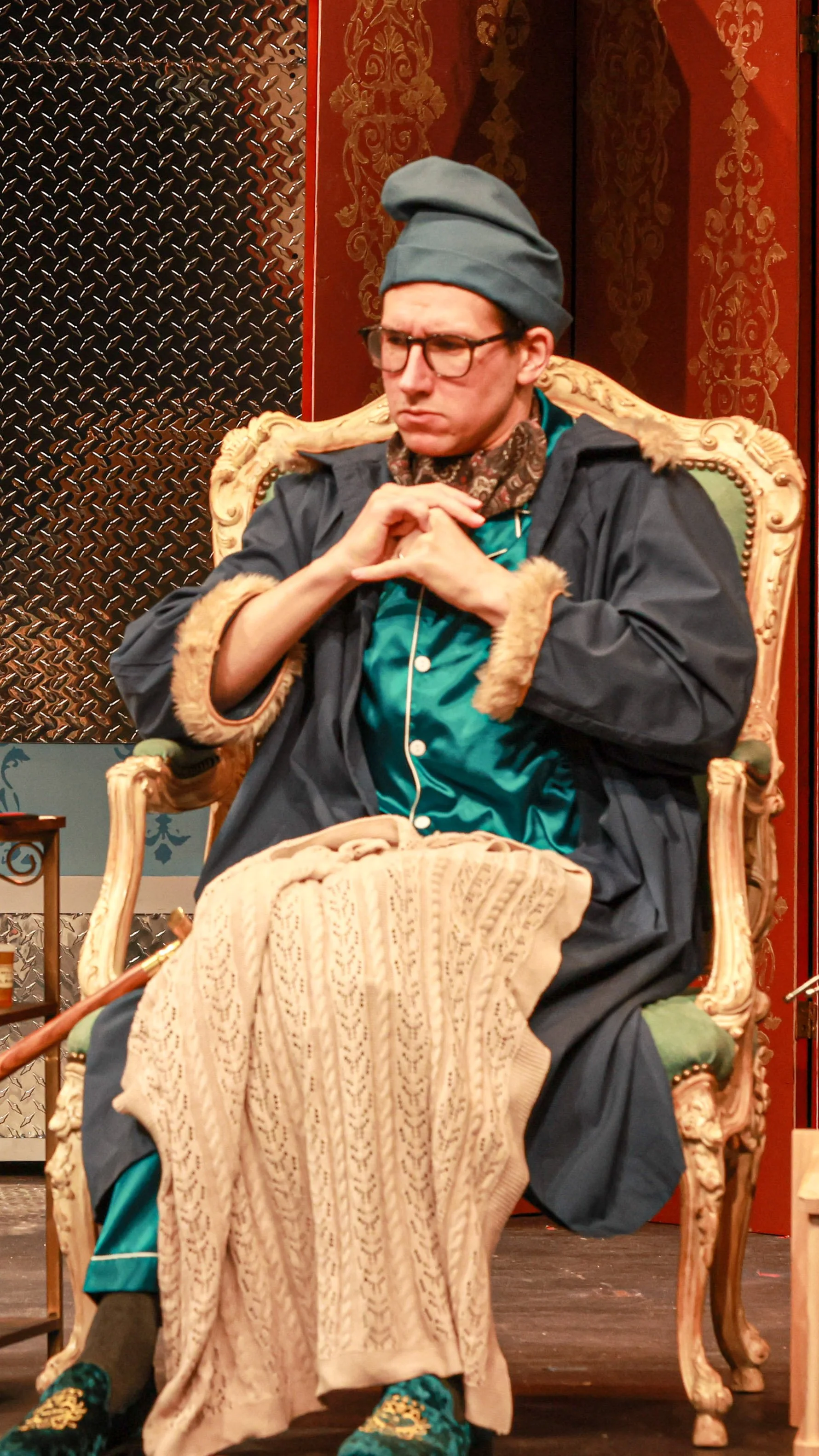 Performer in a cap and robe sits in an ornate chair in Molière’s The Imaginary Invalid.