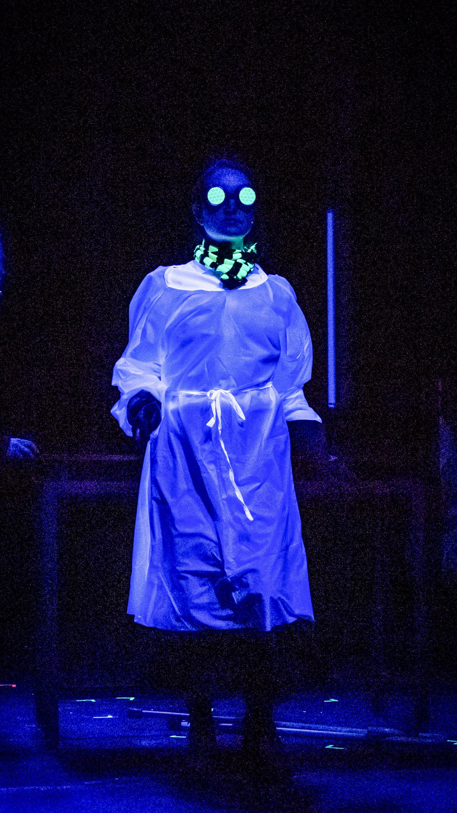 Performer in a white coat and glowing protective mask stands under blue light in Radium Girls.