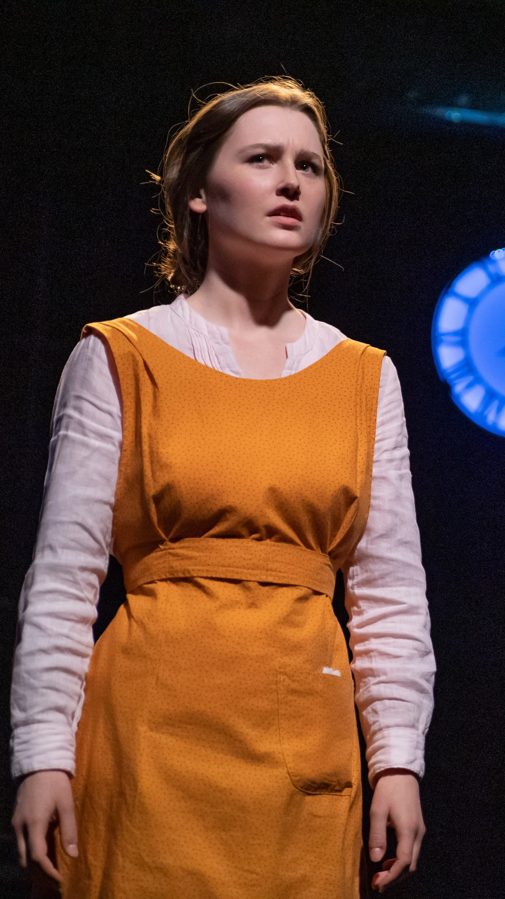 Performer in an orange dress stands under cool blue light in Radium Girls.