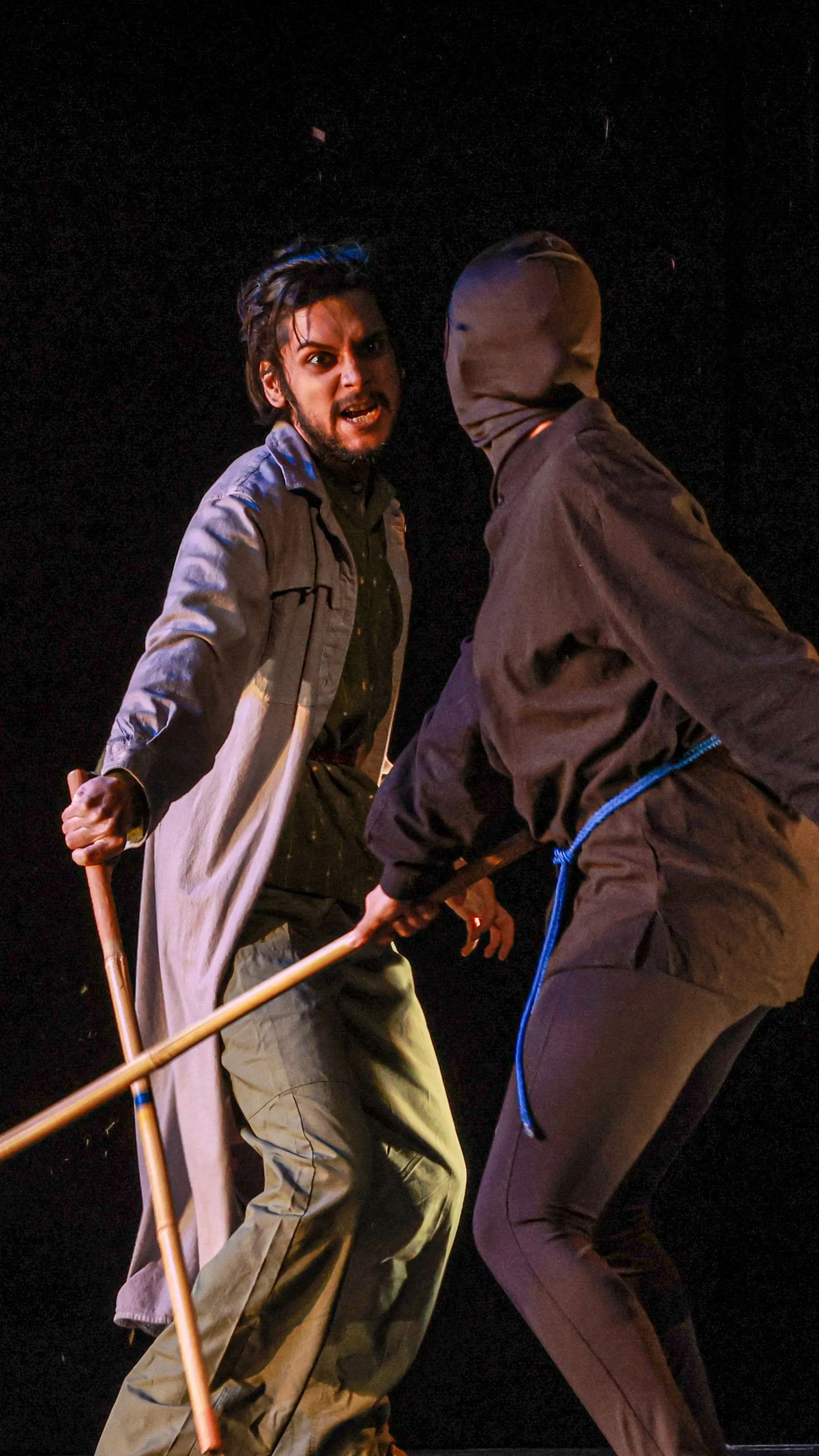 Two performers wield staffs in a close confrontation in LEAR.