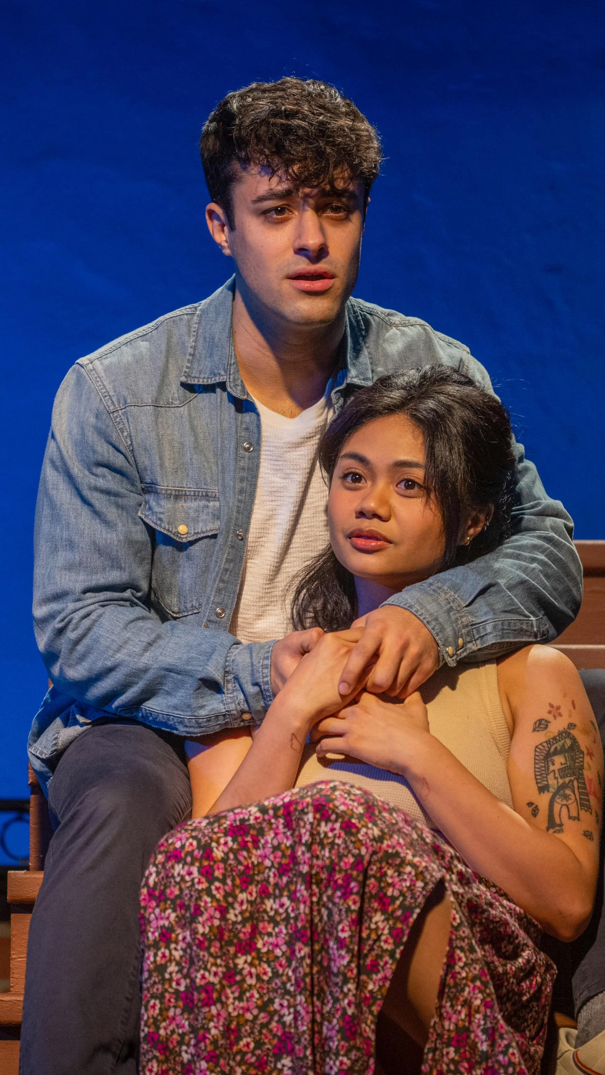 Two seated performers lean together, one with an arm around the other, in Stoop Songs.