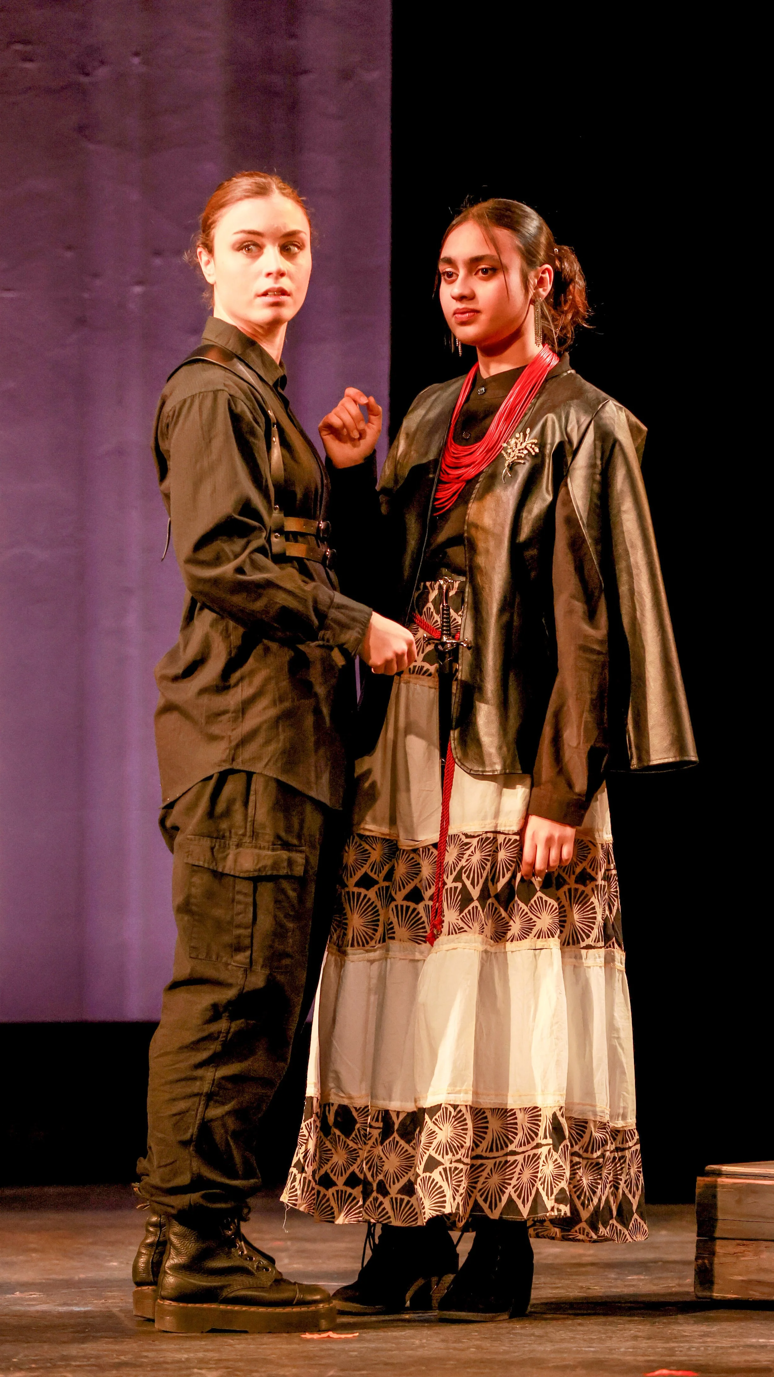 Two performers in dark coats stand together, one clutching a red scarf, in LEAR.