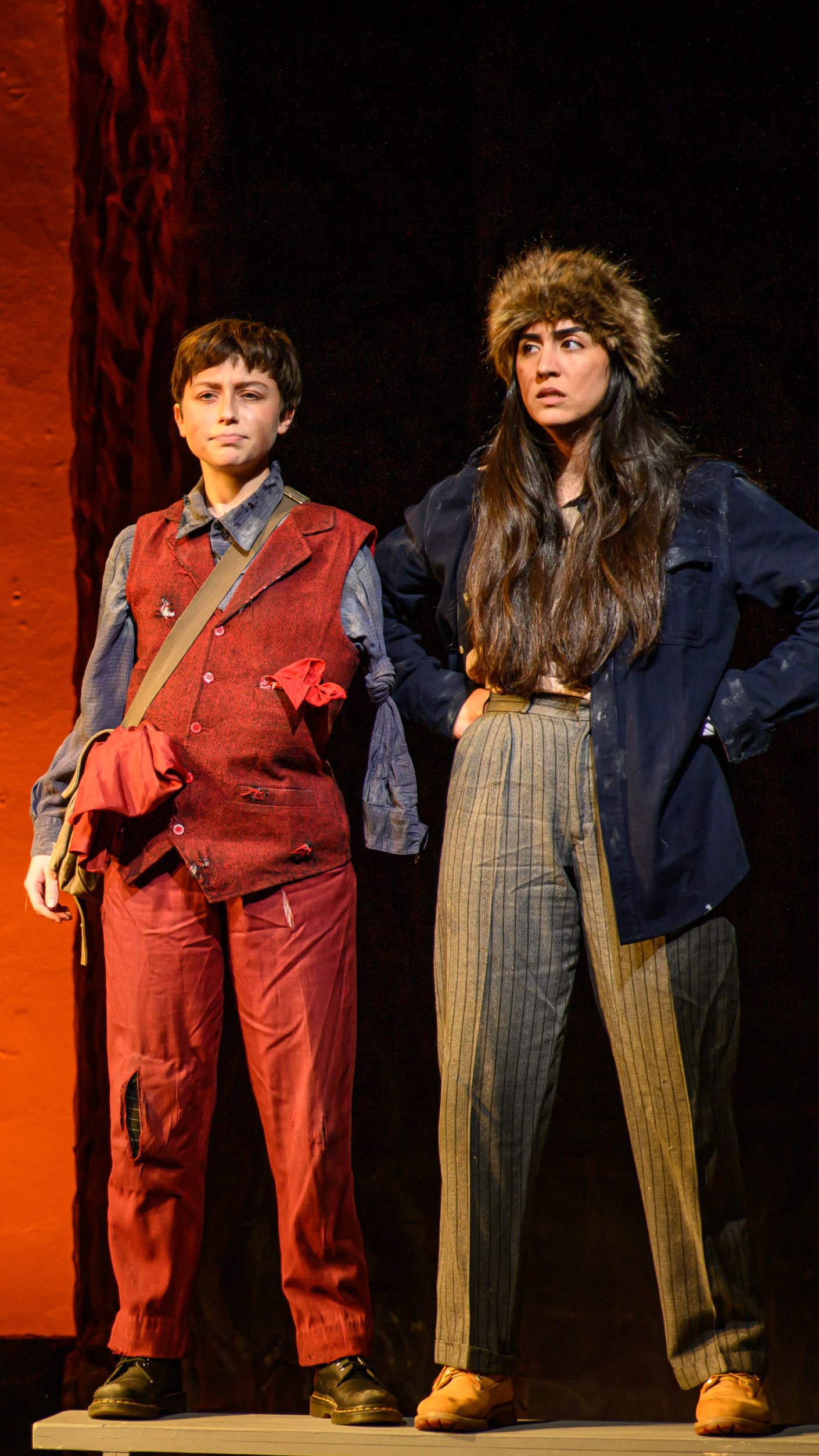 Two performers stand side by side in red and dark expedition costumes in Men on Boats.