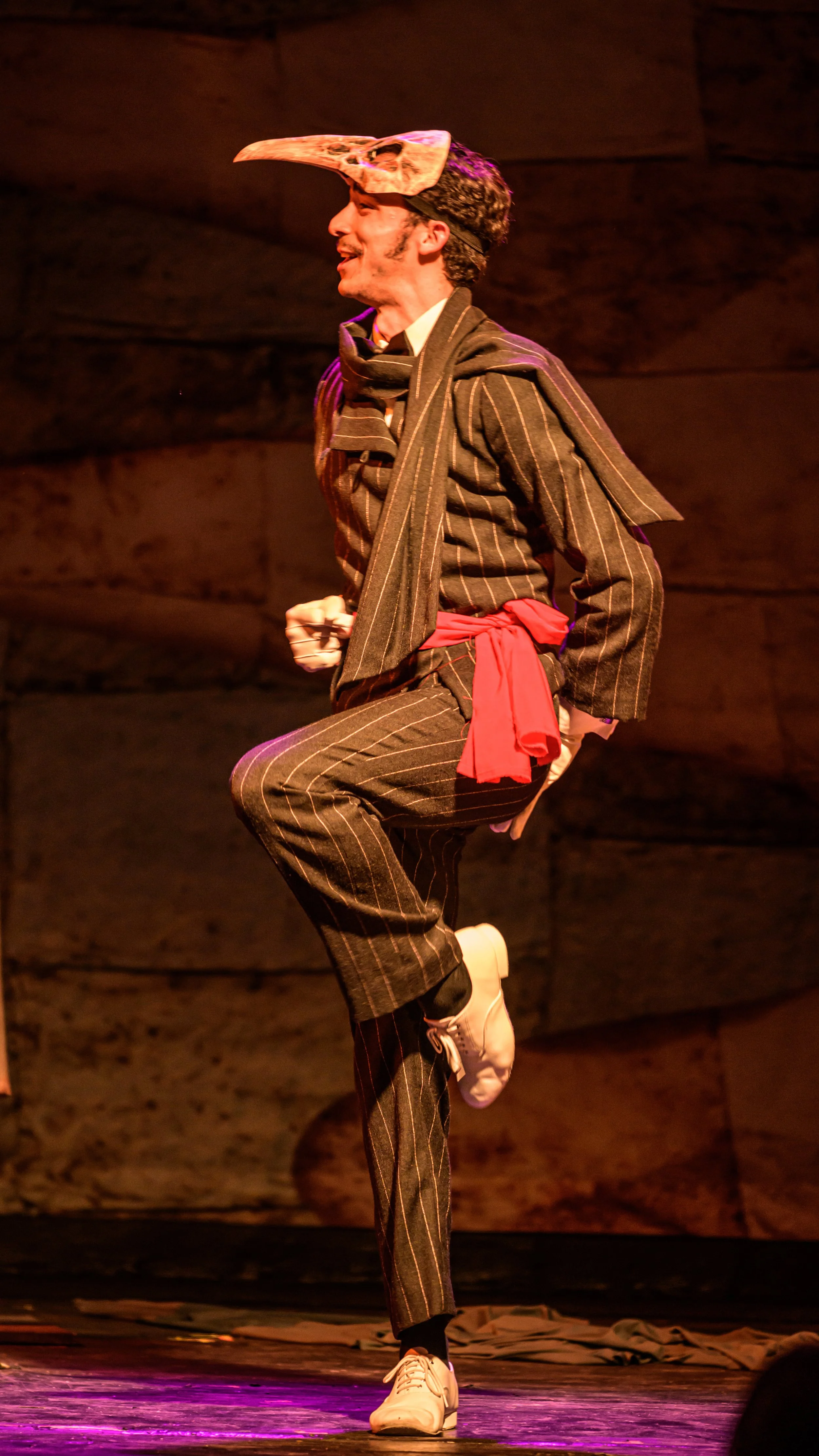 Performer in a striped suit and sculpted cap crosses the stage mid-step in Mr. Burns.