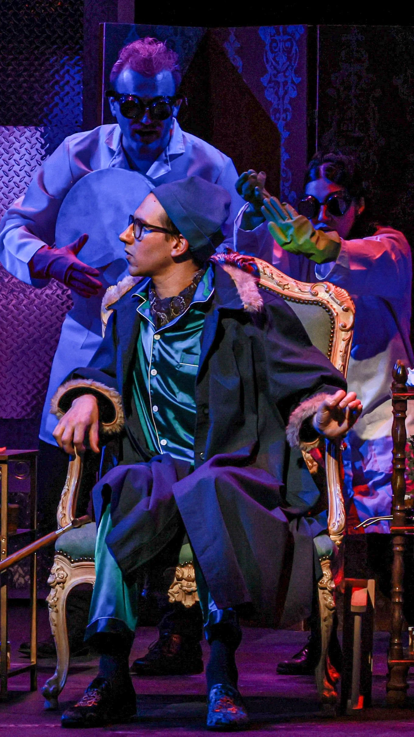Seated performer in sunglasses is surrounded by three costumed figures in Molière’s The Imaginary Invalid.