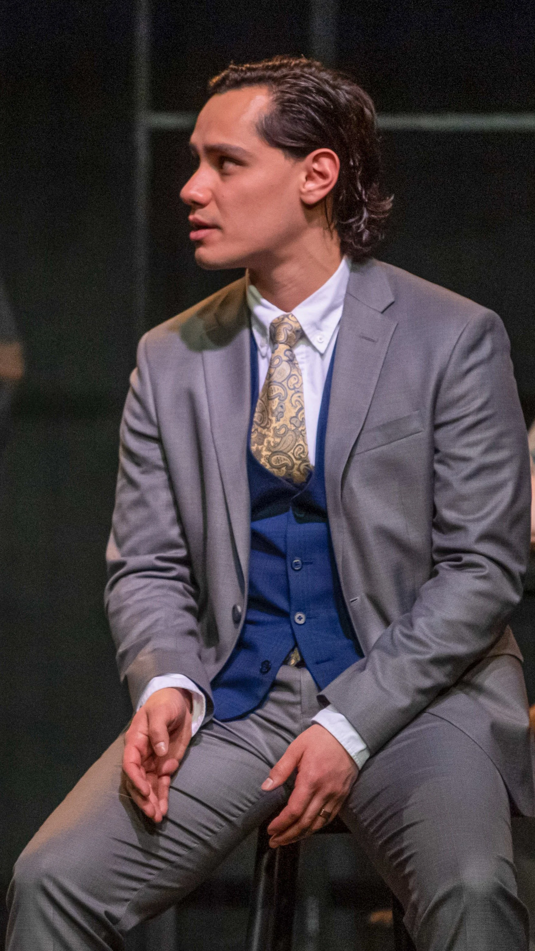 Performer in a gray suit and blue vest sits in profile in Radium Girls.
