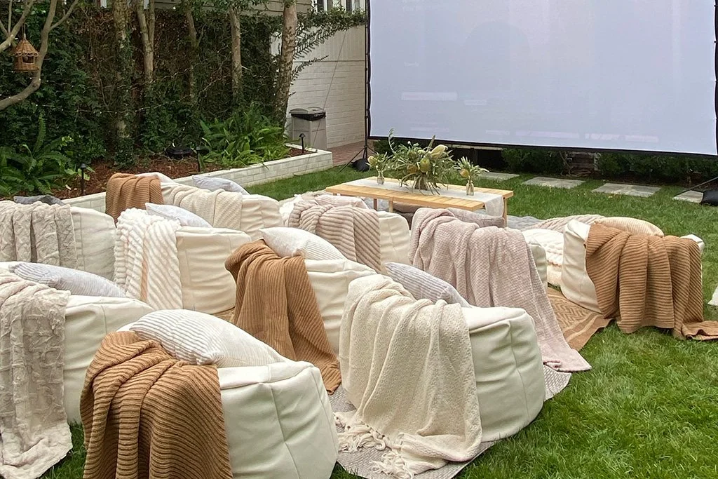 Backyard movie screen with cream lounge seating, layered throws, and a low wooden table