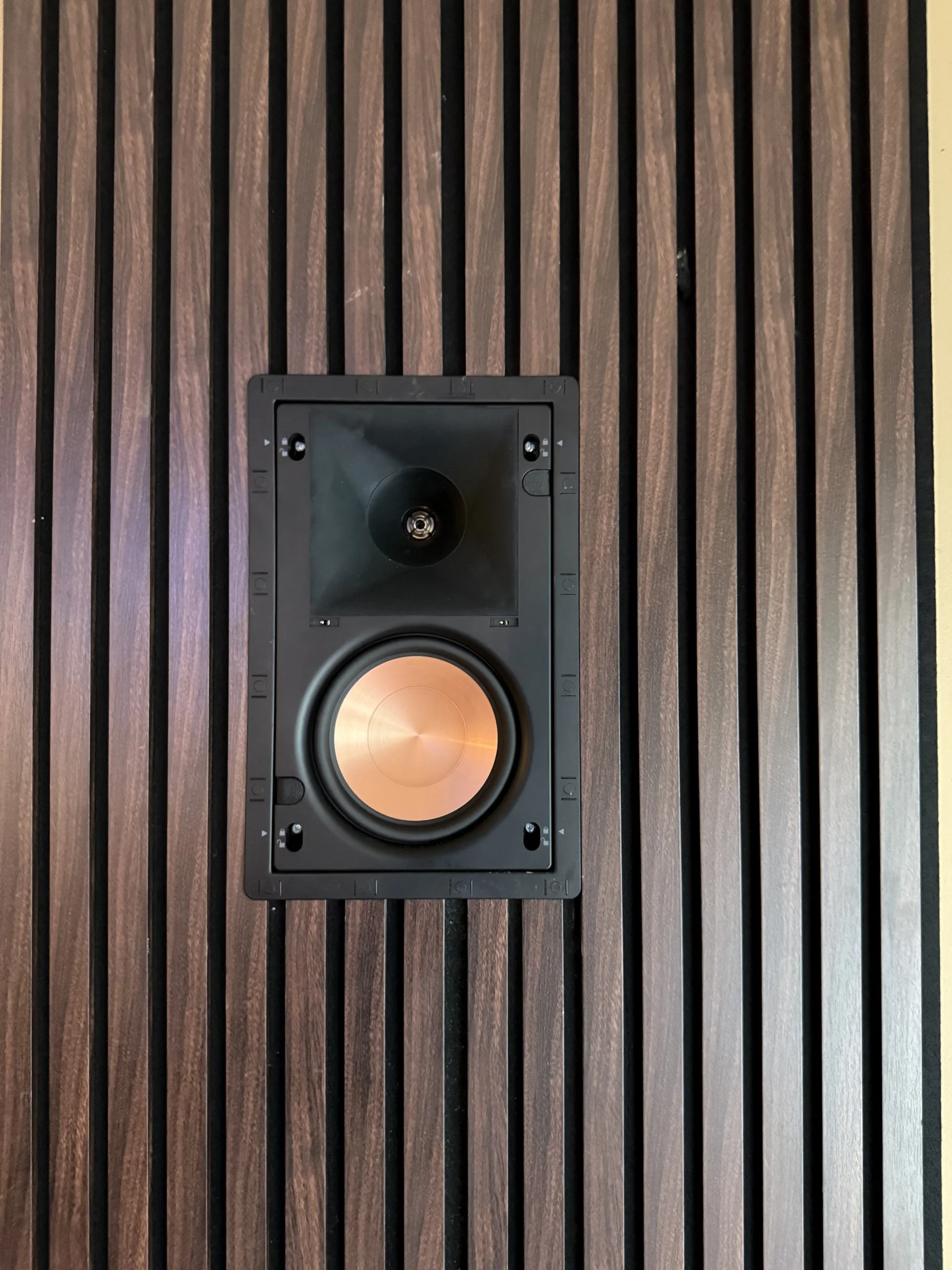 Close-up of an in-wall speaker with its grille removed, showing a copper-colored cone in the slat wall