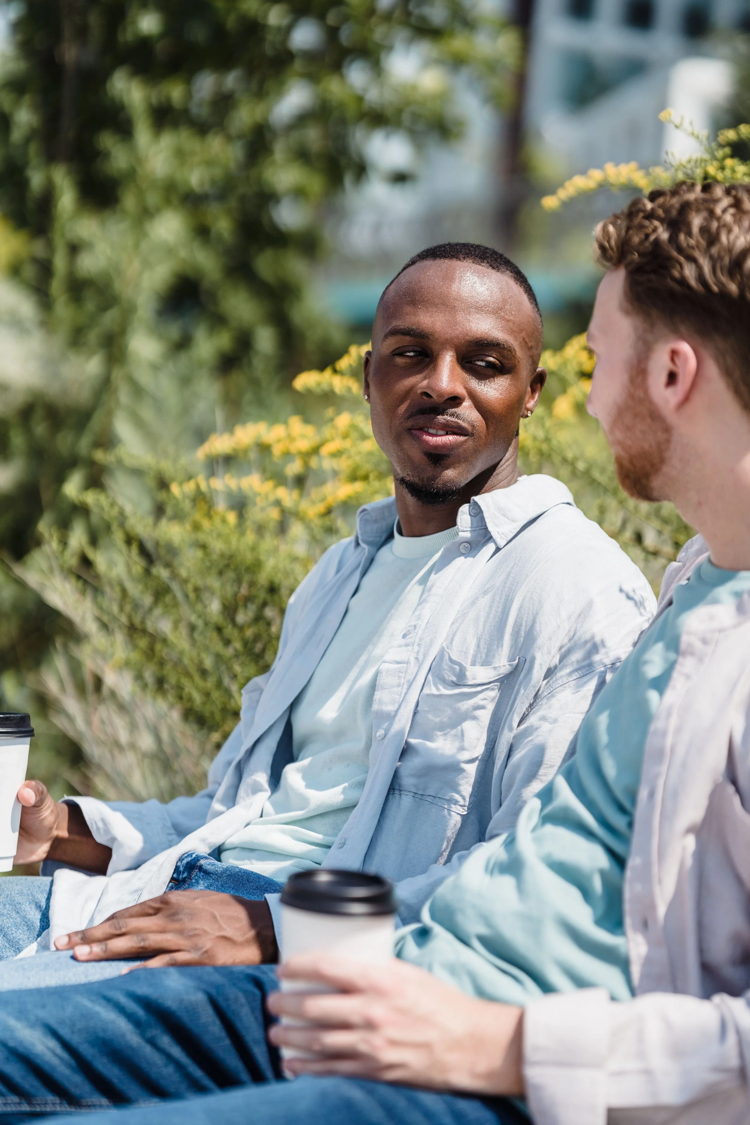 Two men sitting outdoors in a park, talking over coffee.