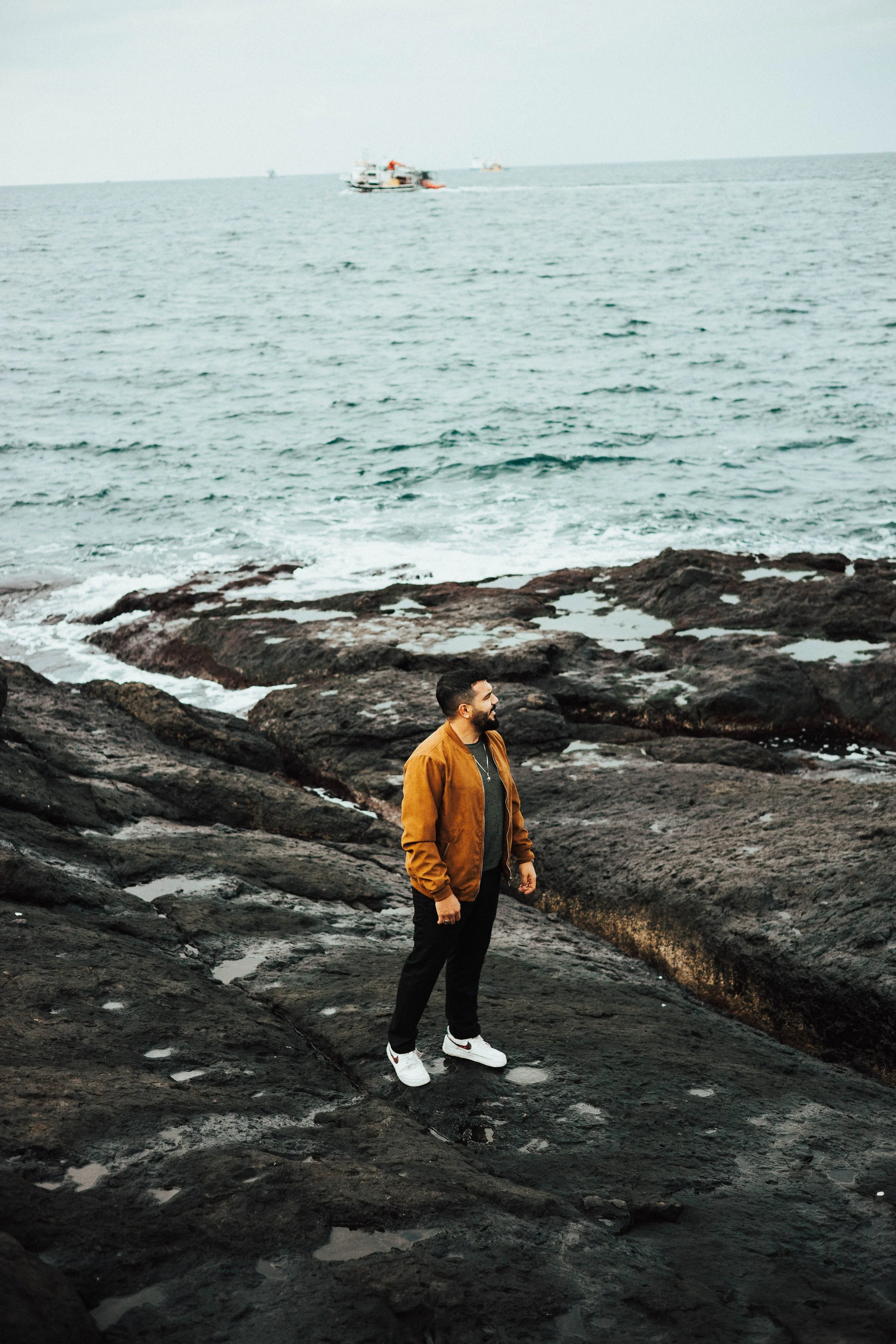 A man in a brown jacket standing on a rocky shore, looking out to sea.