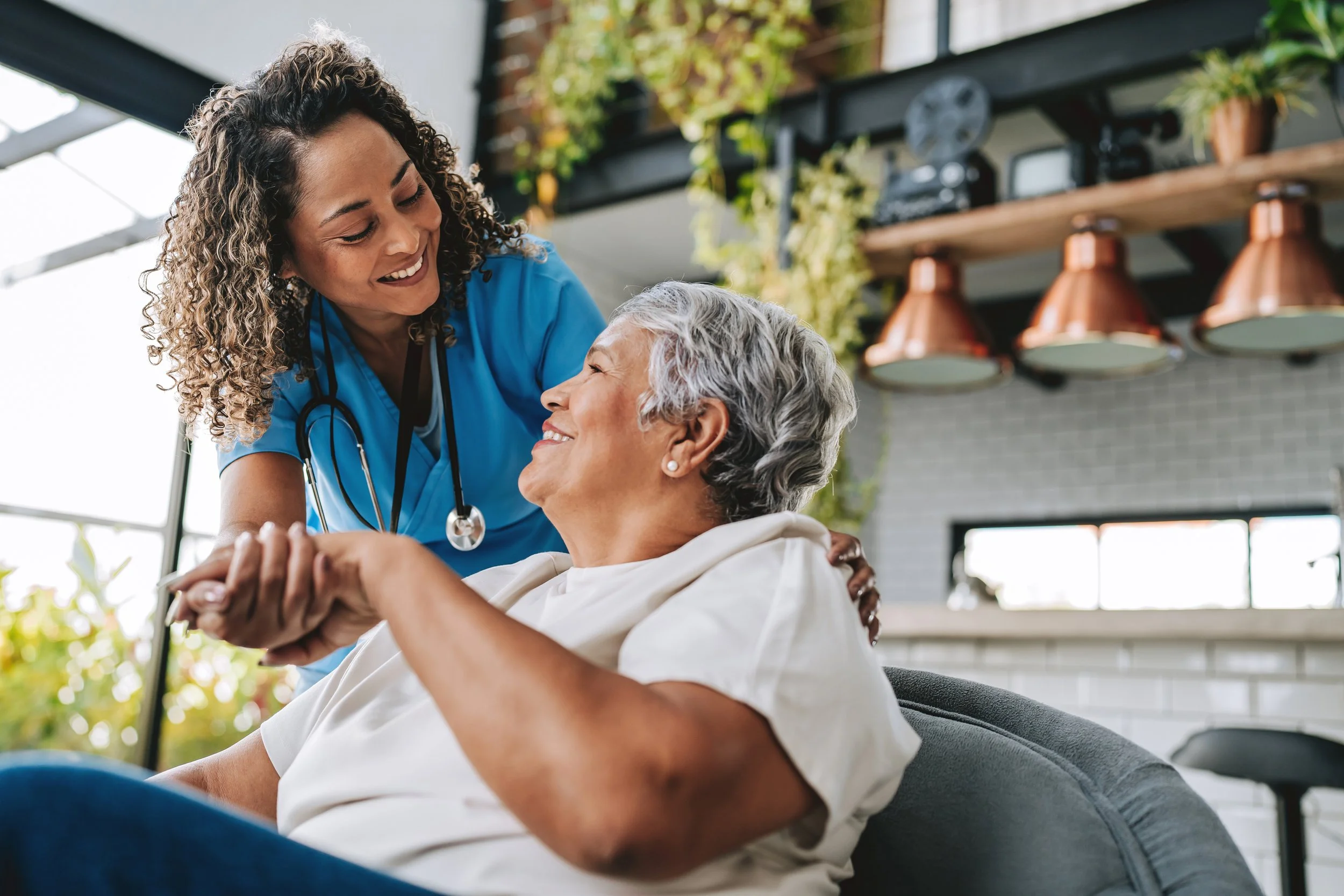 A private duty nurse, wearing blue scrubs and a stethoscope, is smiling and holding the hands of an elderly woman who is sitting in a chair, with both women looking happily at each other inside a bright room.