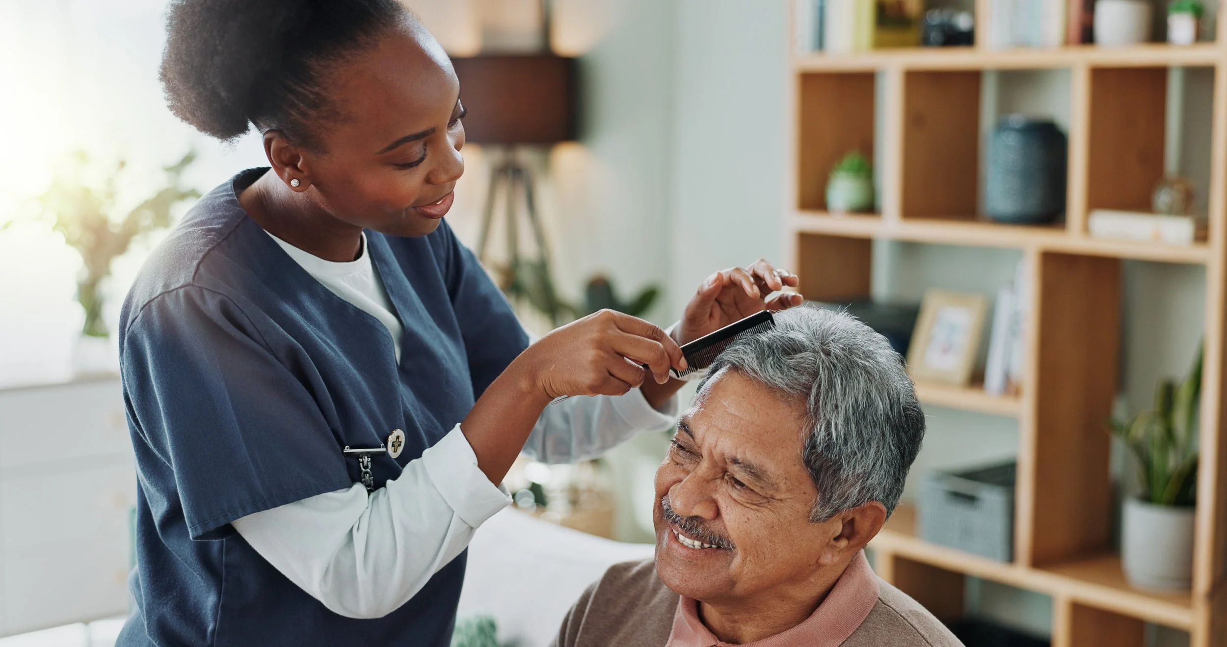 A nurse grooming an elderly man's hair with a comb in a bright room with wooden shelves and plants.