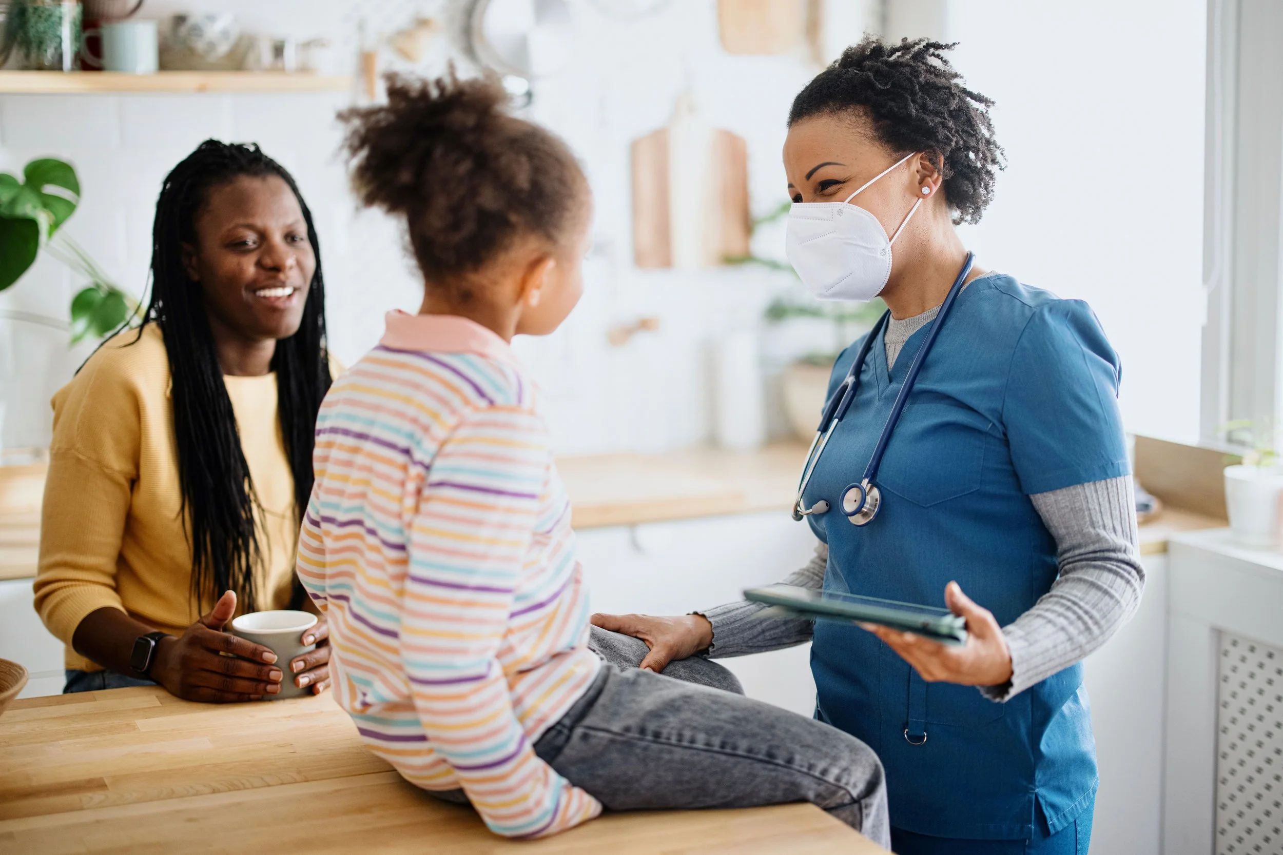 A pediatric nurse wearing a mask talking to a young girl sitting on a counter, with an woman smiling holding a mug nearby.