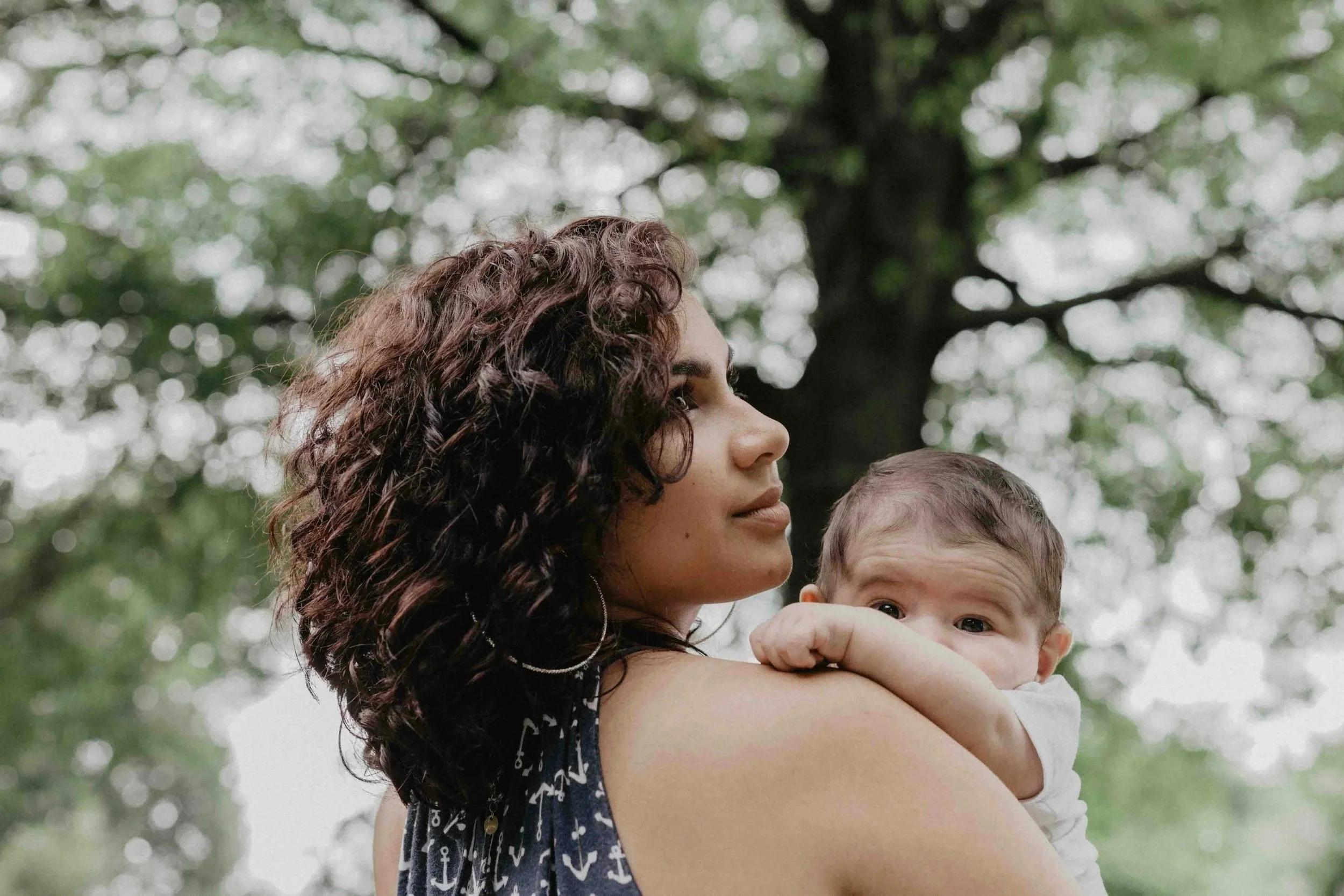 A mother holding her baby on her shoulder beneath a tree