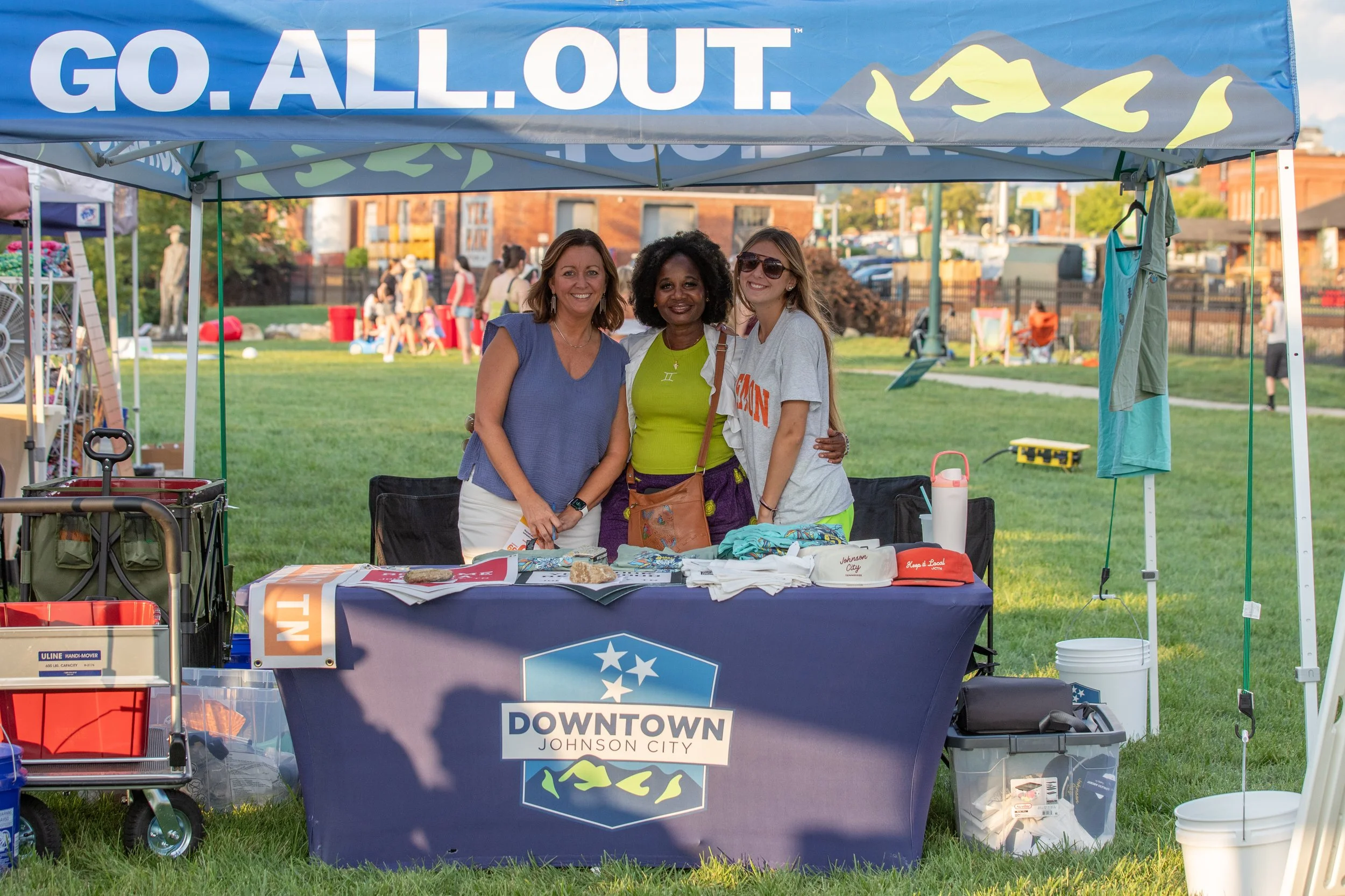 Three volunteers smiling behind the Downtown Johnson City booth at an outdoor event
