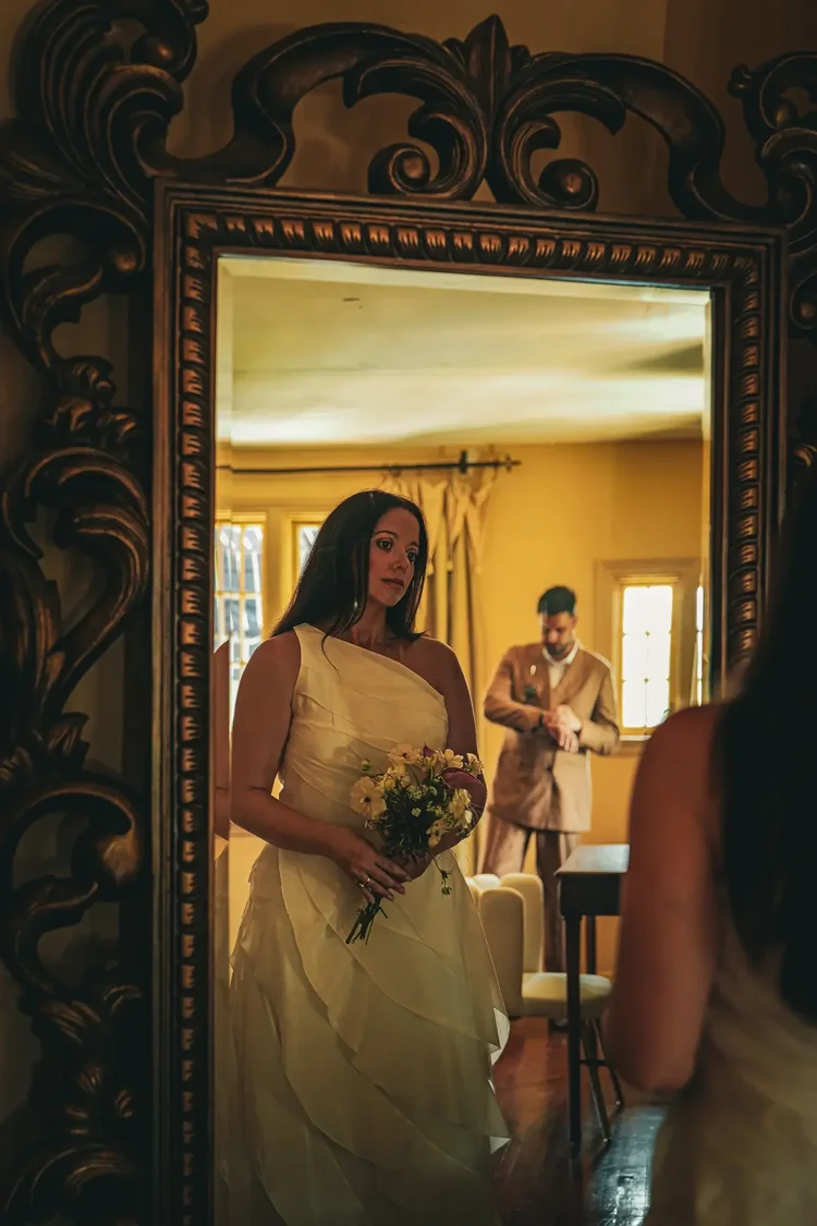 Bride seen in an ornate gilded mirror while getting ready at the Young Mansion, groom in the background