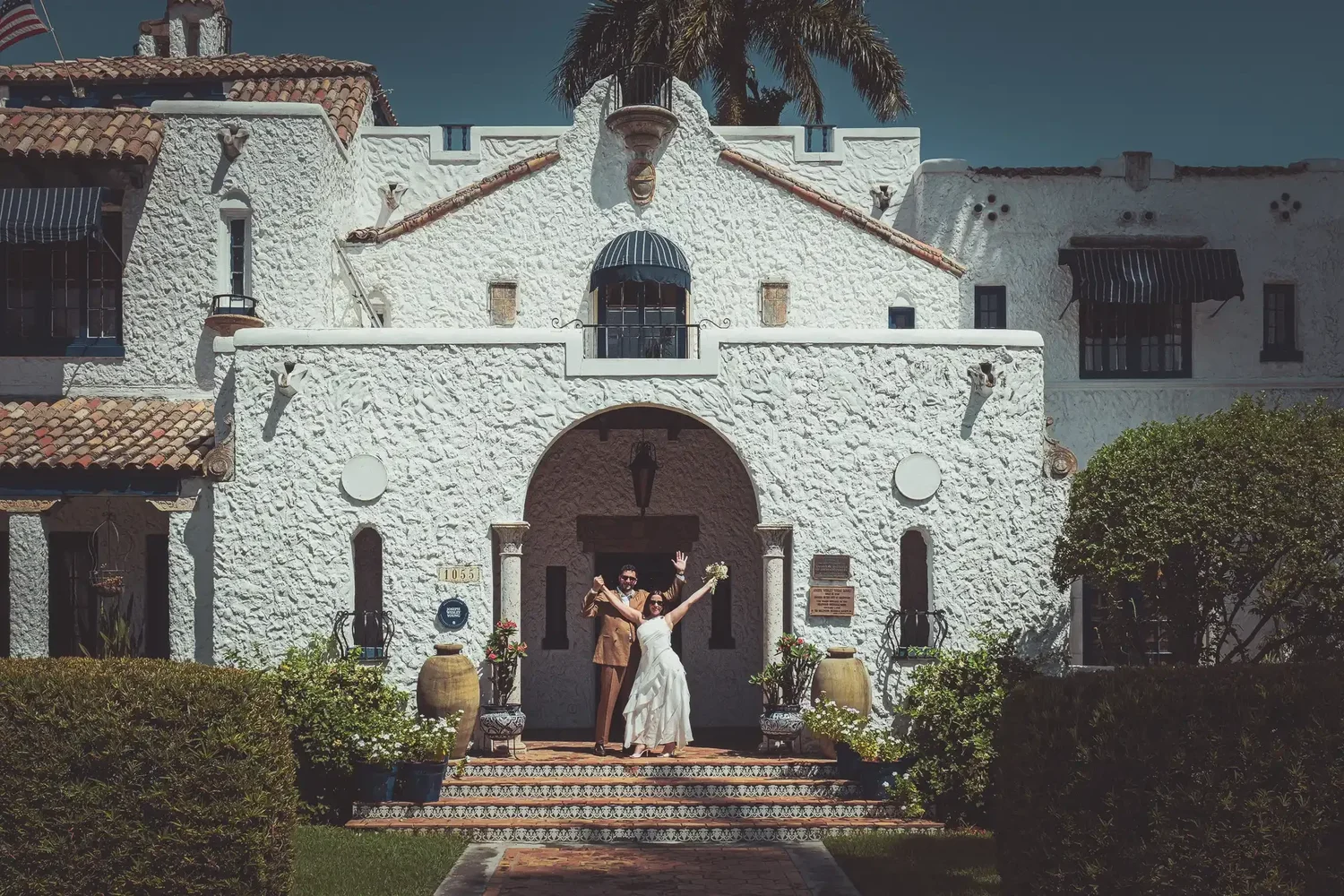 Newlyweds Simon and Laura celebrating with arms raised on the tiled steps of the Young Mansion, a Mediterranean Revival villa in Hollywood, Florida