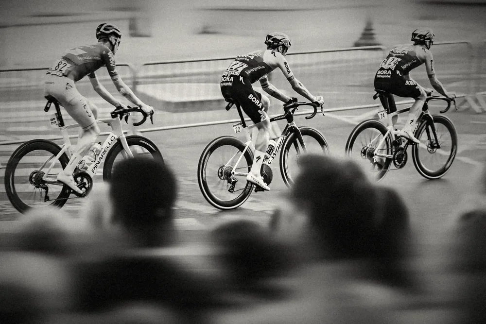Three riders in a line past the crowd, sharp against a blurred Paris, black and white