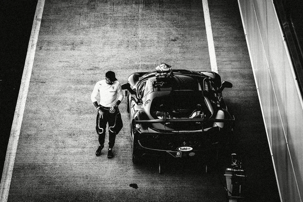 A driver leaning on his race car in the pit lane, high-contrast black and white
