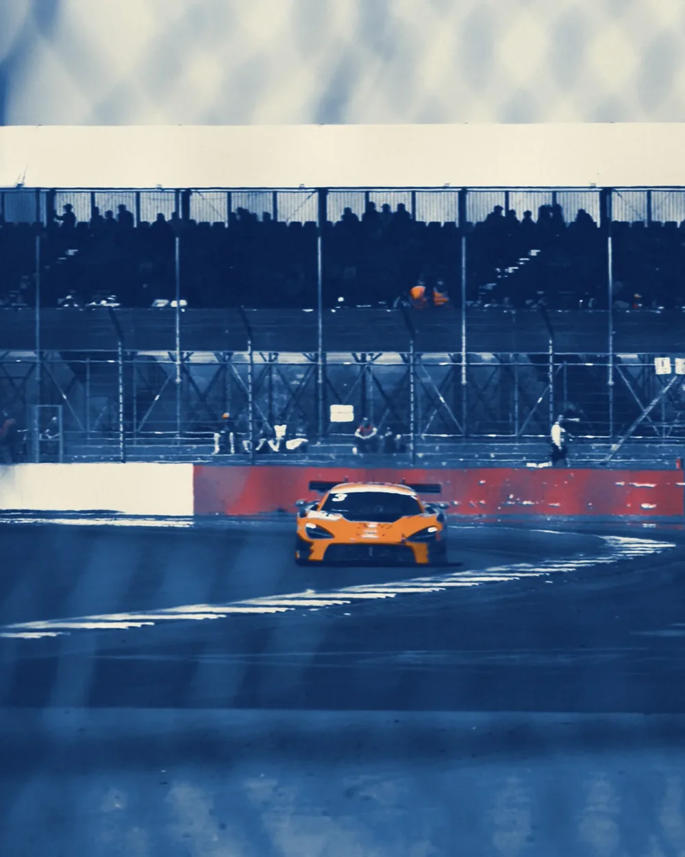 An orange race car heading for the camera under a packed grandstand, cyanotype blue with orange overprinted