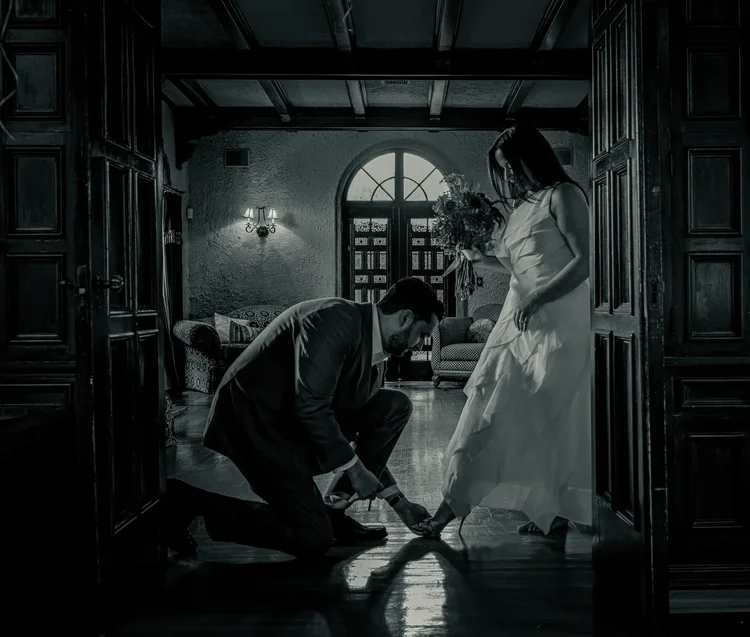 Groom kneeling to fasten the bride's shoe in a dark wood-panelled hall at the Young Mansion, black and white