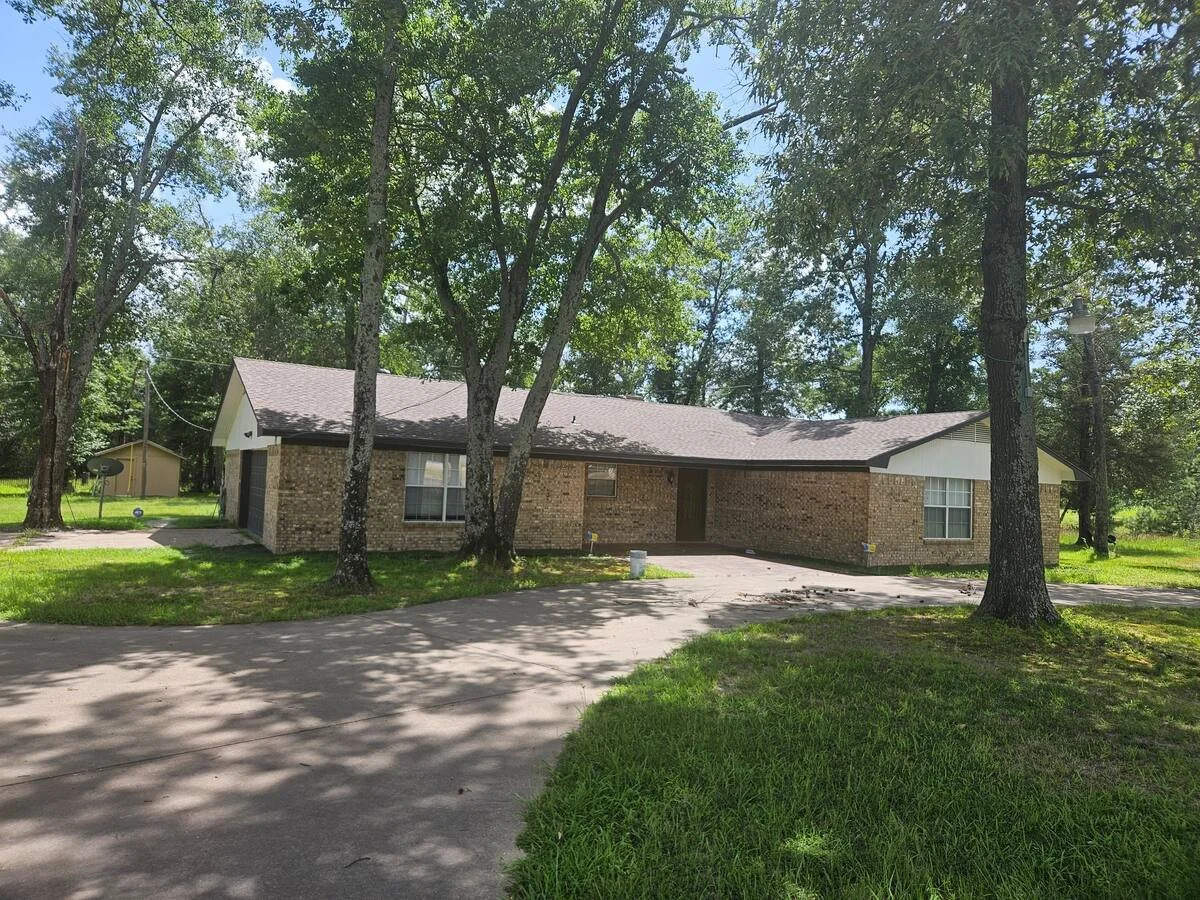 Exterior view of Rancho 5 at Pinoy Ranch in Wills Point, Texas. A single-story brick ranch home featuring a wide driveway, large shaded lawn with mature trees, and a quiet, wooded surroundings.