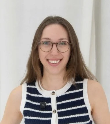 Abbie souriante avec des lunettes, portant un gilet rayé marine et blanc, debout devant un rideau blanc.
