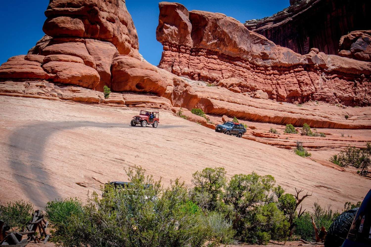 Red open-top FJ40 and a dark Land Cruiser wagon on a slickrock slope under red fins on the Seven Mile Rim trail near Moab, Utah