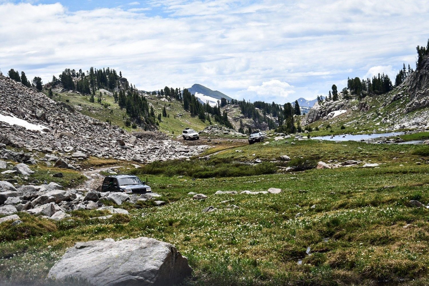 Trucks crossing a wide rocky alpine basin with snowfields and peaks near Cooke City, Montana