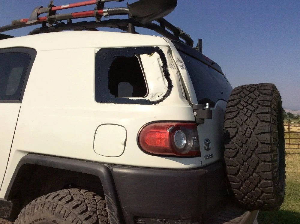 Close view of a white FJ Cruiser with a shattered rear quarter window on a rocky forest trail in Montana