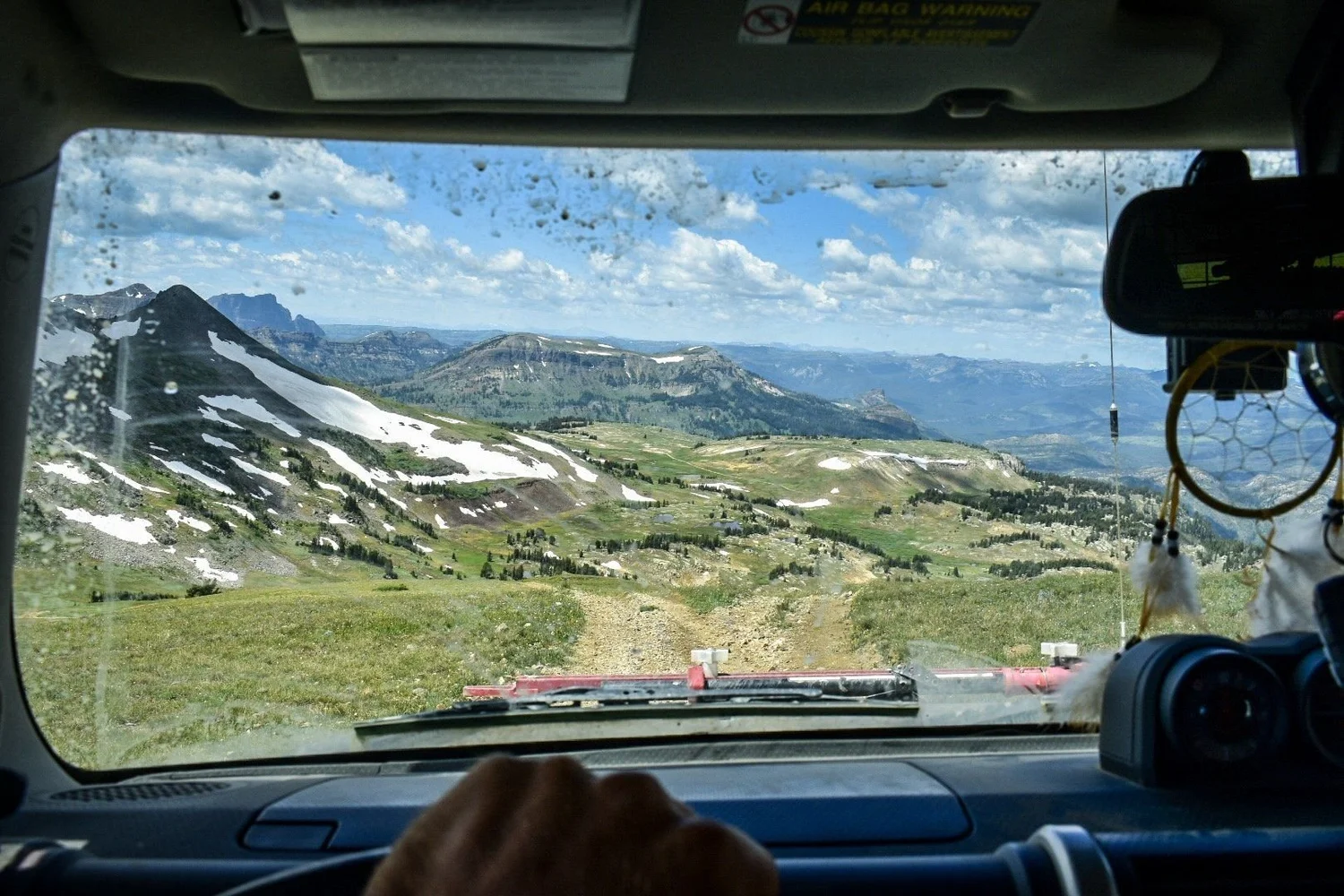 View through a dusty windshield of a two-track crossing high tundra with snowfields and a distant valley near Cooke City, Montana