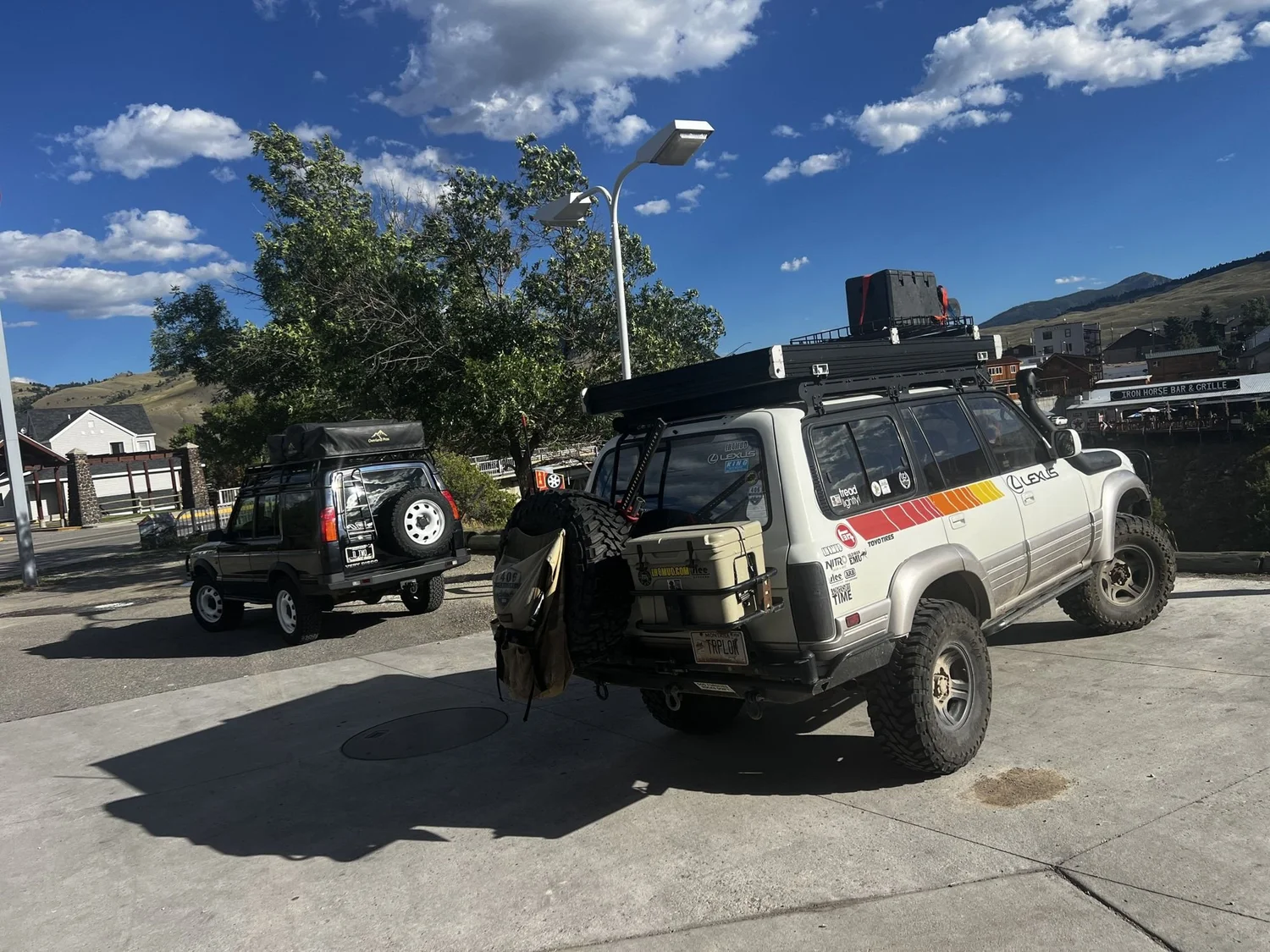 Striped Lexus LX450 with a roof rack and open rear door parked in a sunny lot next to another Land Cruiser near Cooke City, Montana