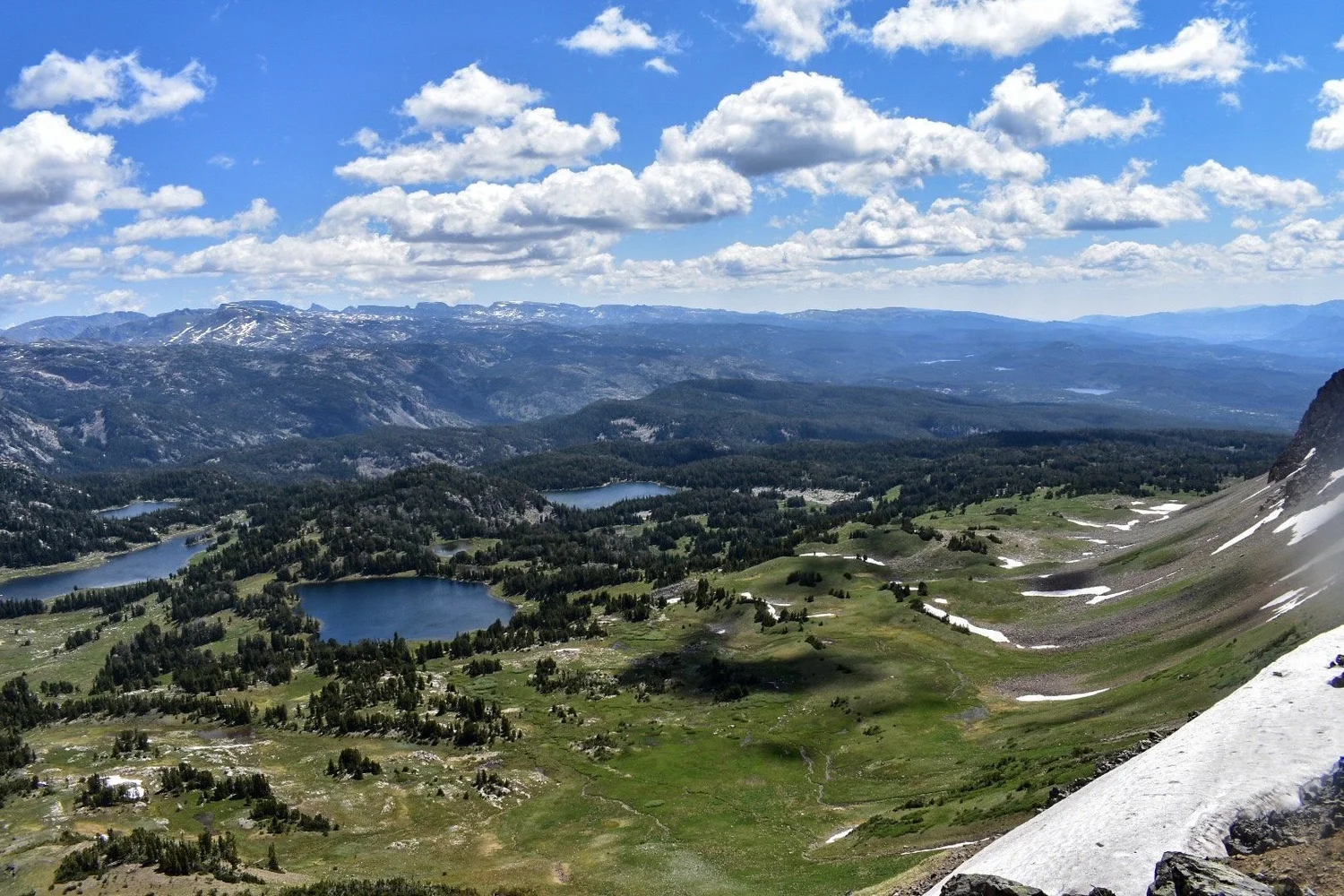 High overlook of a green basin dotted with blue lakes and forest, with a snowfield in the foreground near Cooke City, Montana