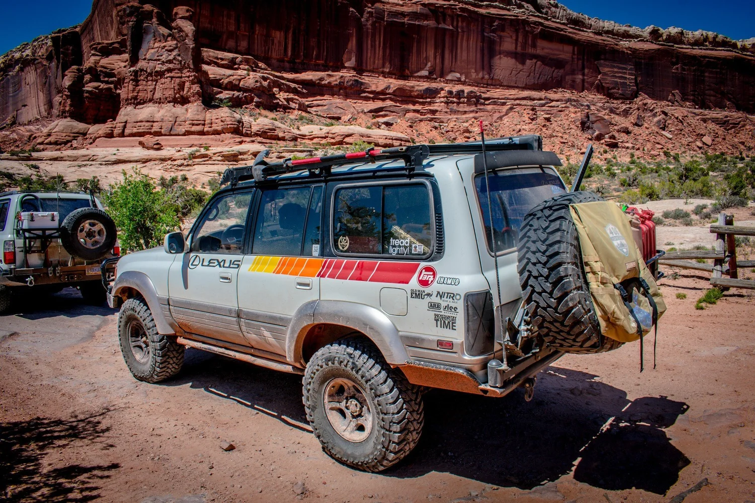 Rear quarter of a silver Lexus LX450 with a retro Lexus stripe and swing-out tire carrier below red cliffs on the Seven Mile Rim trail near Moab, Utah