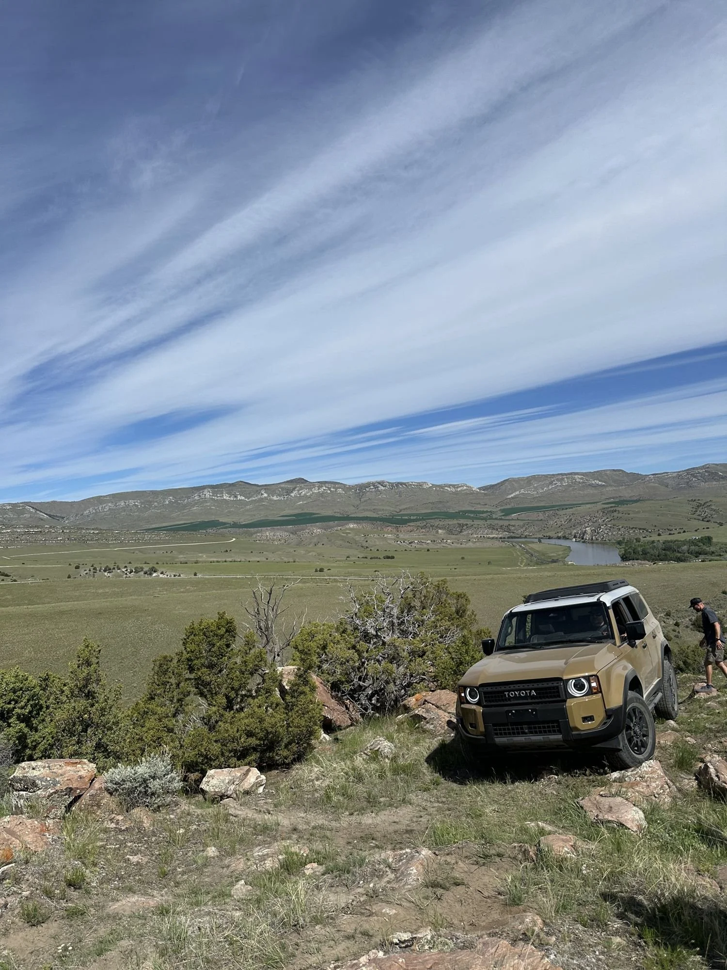Tan Land Cruiser 250 parked on a rocky ridge above a wide green valley under streaky clouds at XOverland Ranch near Bozeman, Montana