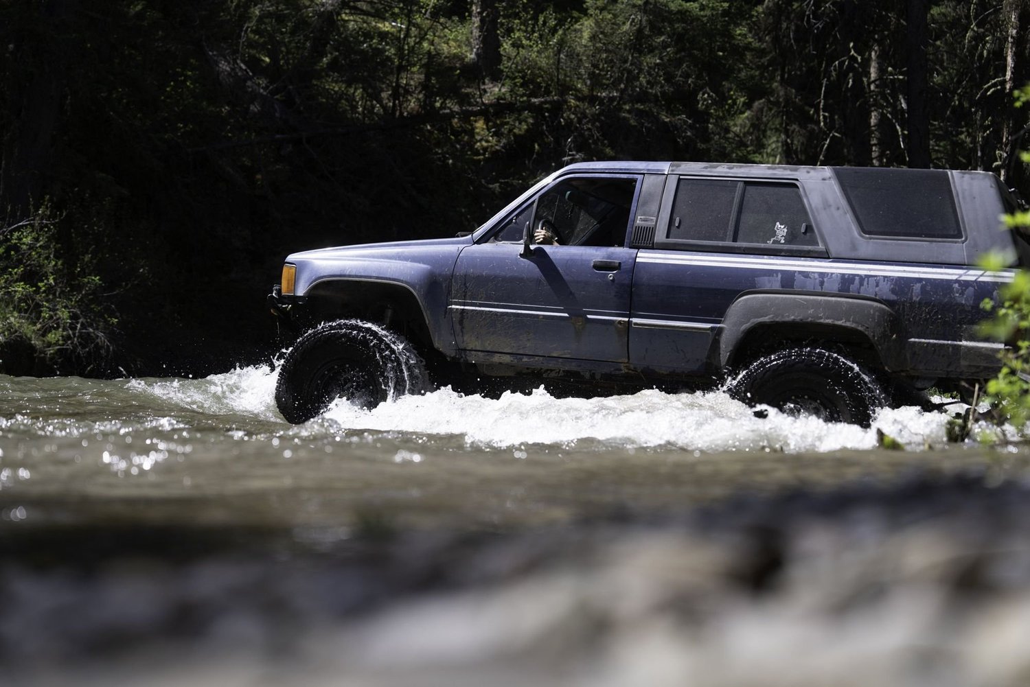 Blue first-generation Toyota 4Runner crossing a river with water spraying from its tires on the Middle Fork Judith River road in the Little Belt Mountains, Montana
