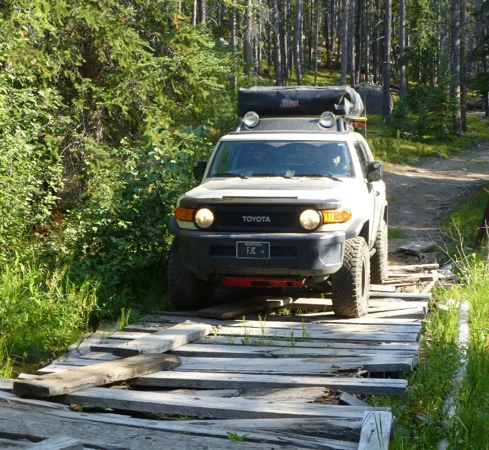 Silver FJ Cruiser crossing a weathered, broken plank bridge in the forest