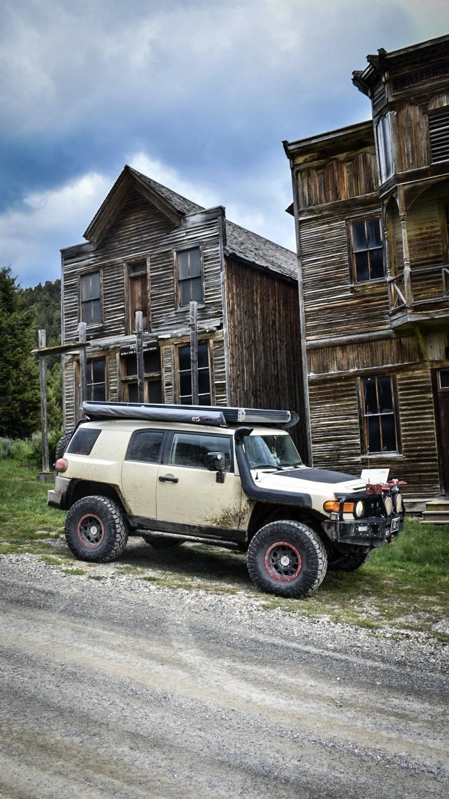 Tall weathered Fraternity Hall with a balcony beside another wooden building, a tan FJ Cruiser parked in front in the Elkhorn ghost town near Boulder, Montana