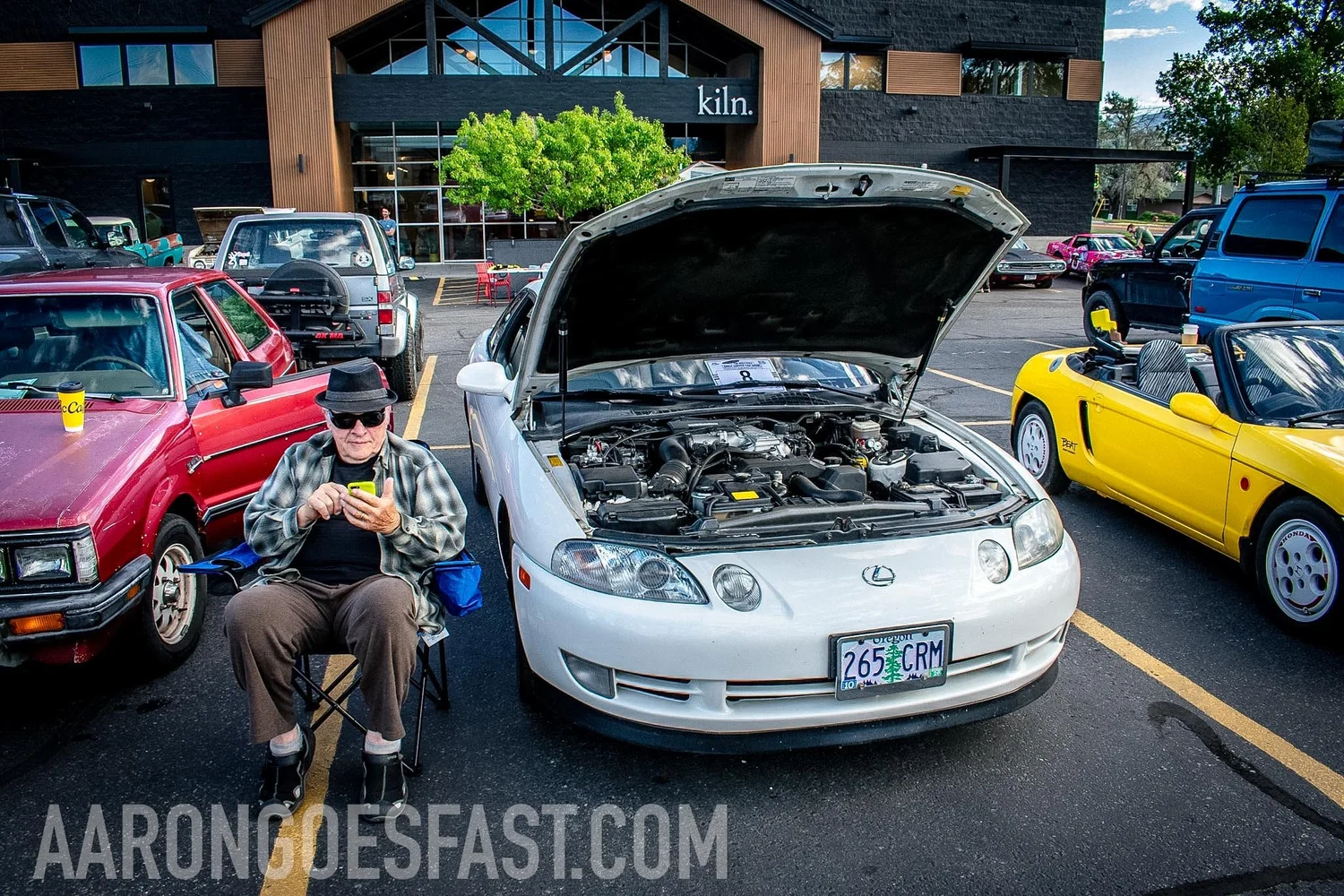 White Lexus SC400 coupe with the hood open next to a yellow Honda Beat at the Roly-Poly Daily Driver Show in Bozeman