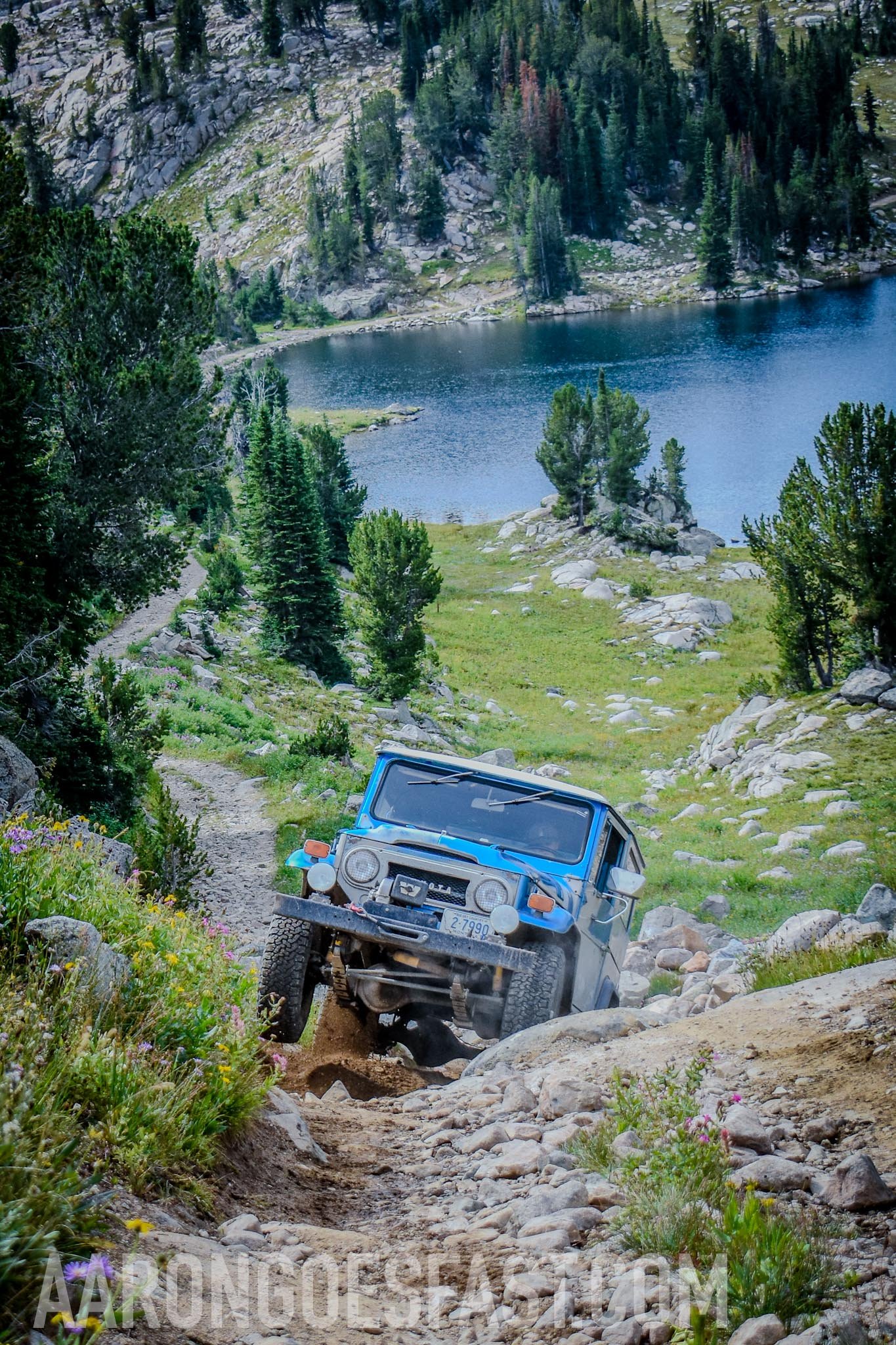 Blue and white Toyota FJ40 Land Cruiser on a rocky trail above an alpine lake on the 406 Cruisers Goose Lake run near Cooke City, Montana