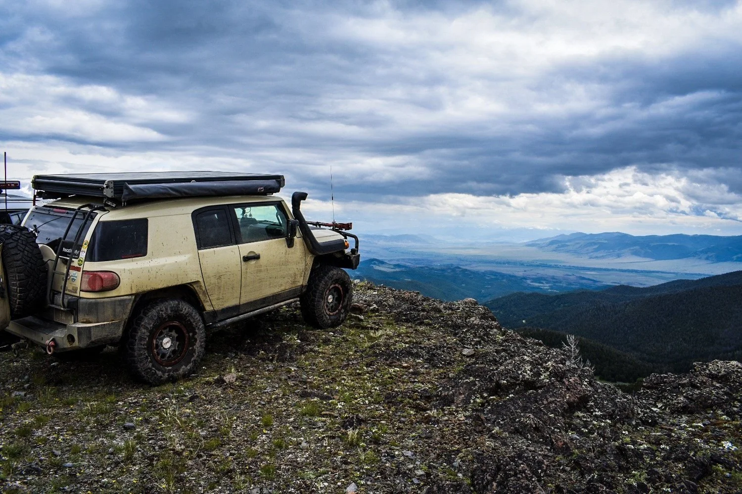 Tan FJ Cruiser parked at the edge of a rocky overlook above valleys and distant mountains under storm clouds in the Elkhorn Mountains near Boulder, Montana