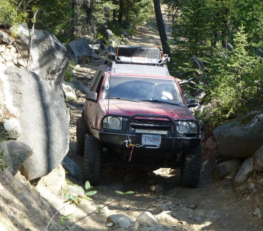 Maroon Nissan Xterra with a winch bumper crawling through a rocky slot in the trees on a rocky forest trail in Montana