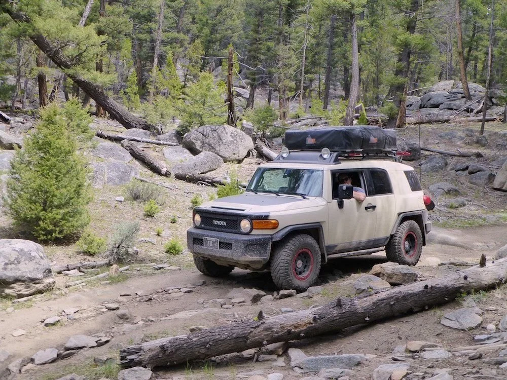 Tan Toyota FJ Cruiser with red wheels and a rooftop tent threading through boulders and downed logs on the Ringing Rocks trail near Pipestone, Montana