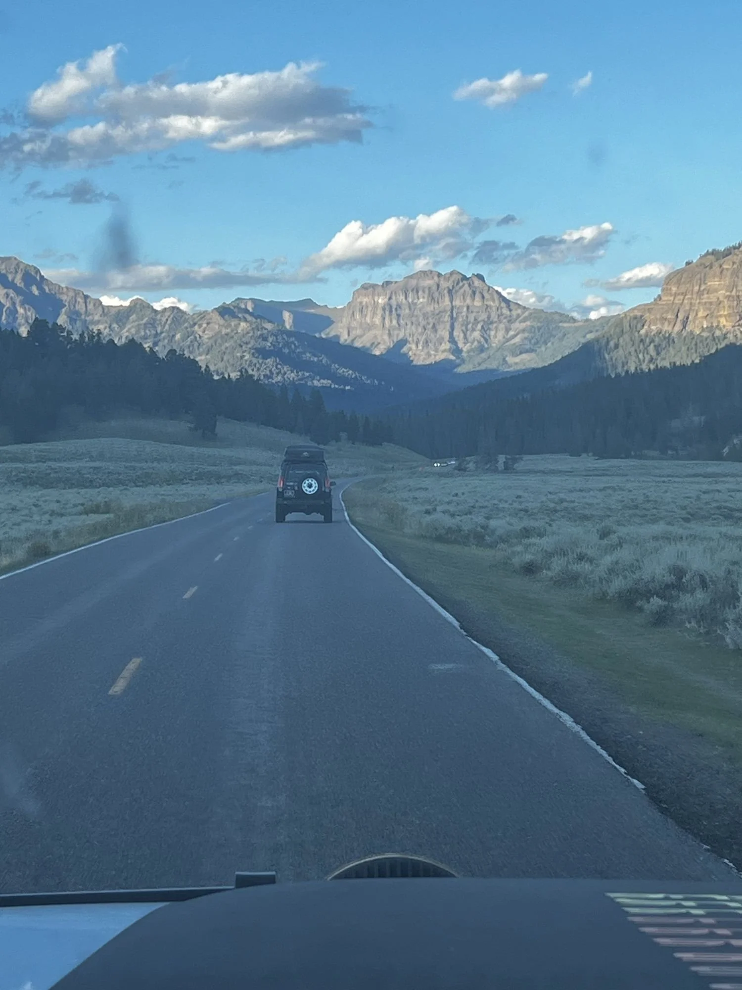 View over a truck hood of a two-lane road heading toward rugged peaks with a truck ahead near Cooke City, Montana