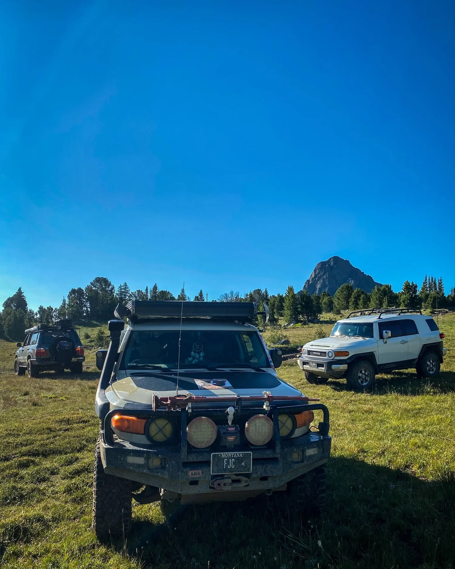 Tan Toyota FJ Cruiser with a steel bumper and a white FJ Cruiser parked in a meadow under a clear blue sky on the Independence road above the Boulder River, Montana